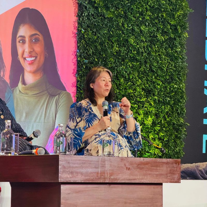 A woman in a blue floral dress speaks into a microphone on a panel, seated at a wooden table with water bottles. A large portrait and greenery backdrop are behind her.