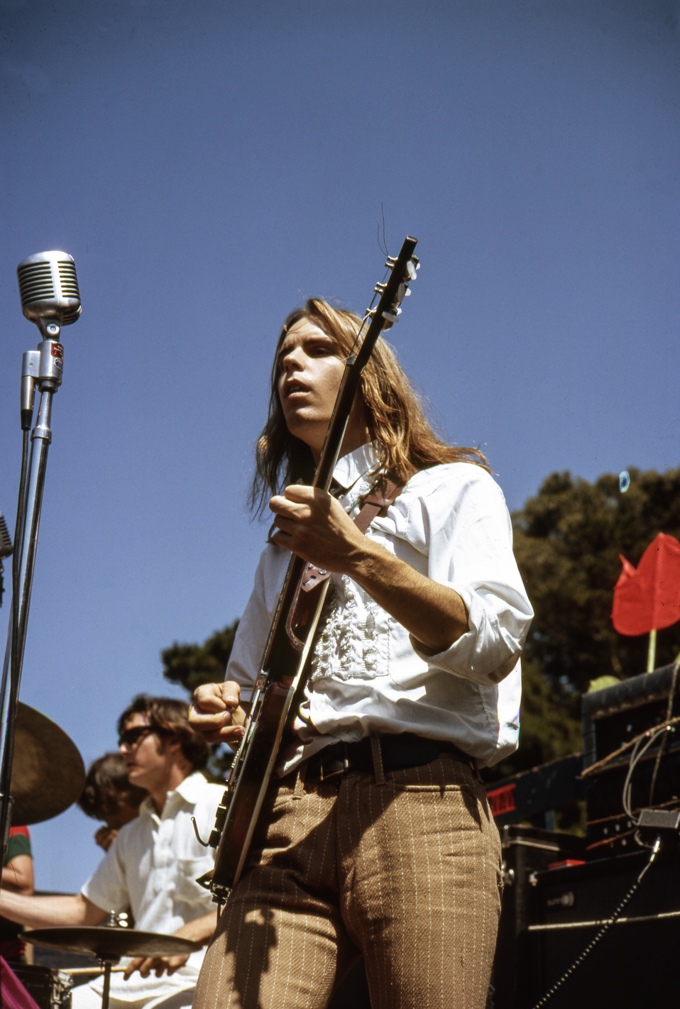 A musician plays electric guitar outdoors, wearing a white shirt and brown pants. A vintage microphone stands nearby, with a drummer in the background.