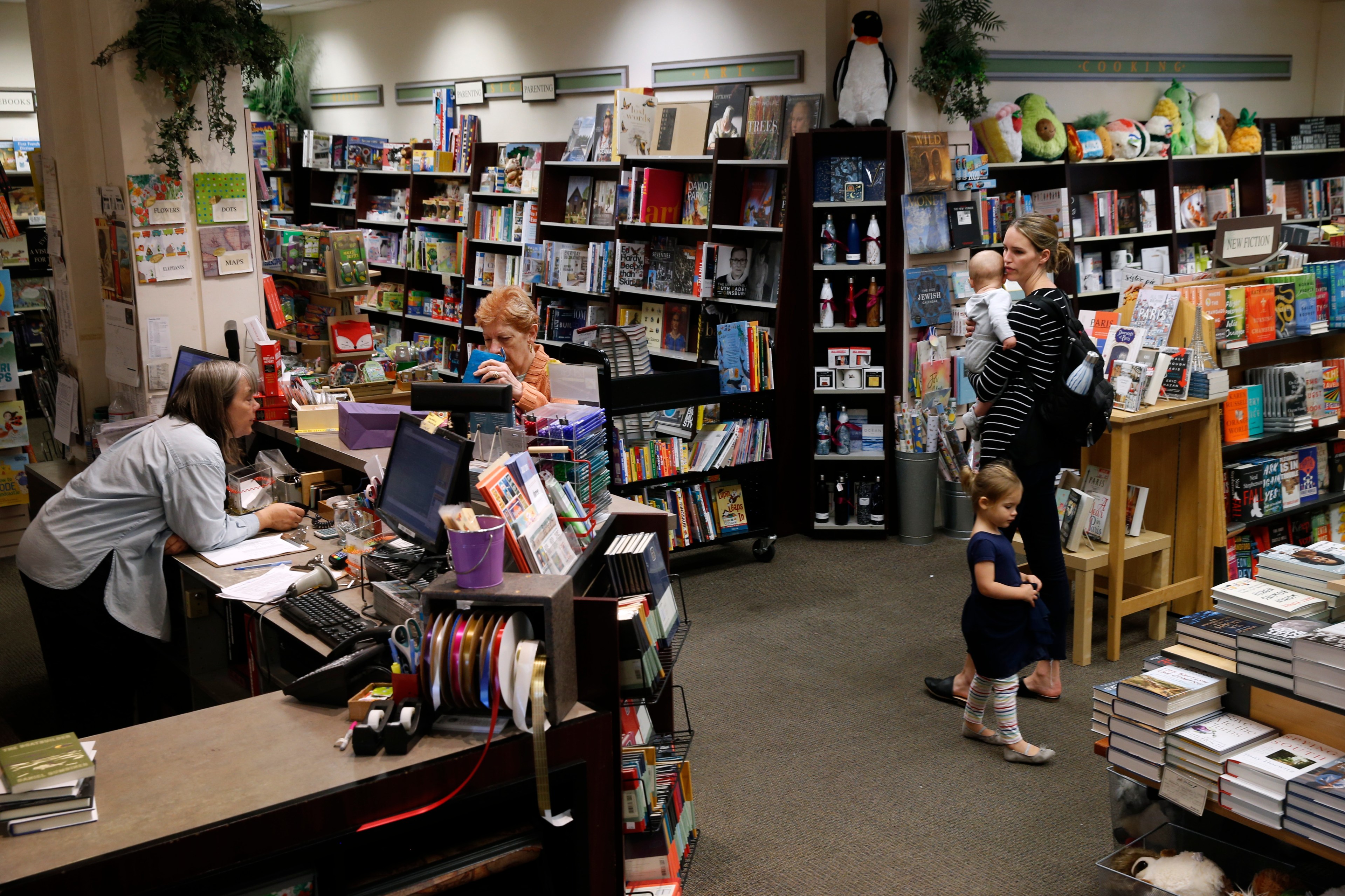 A busy bookstore with shelves full of books, two women at the counter, and a mother with two young children browsing nearby.