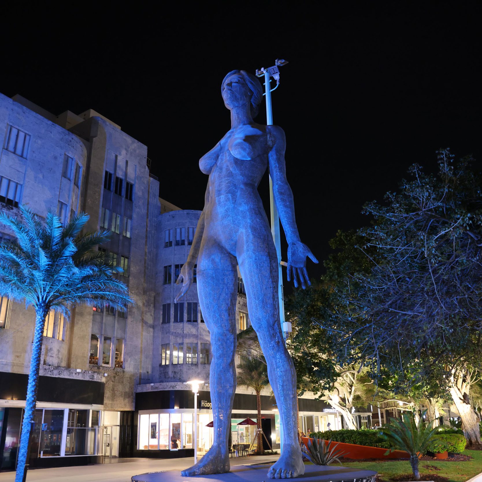 A tall, blue-lit statue of a standing figure is seen at night, set against a backdrop of modern buildings and palm trees.