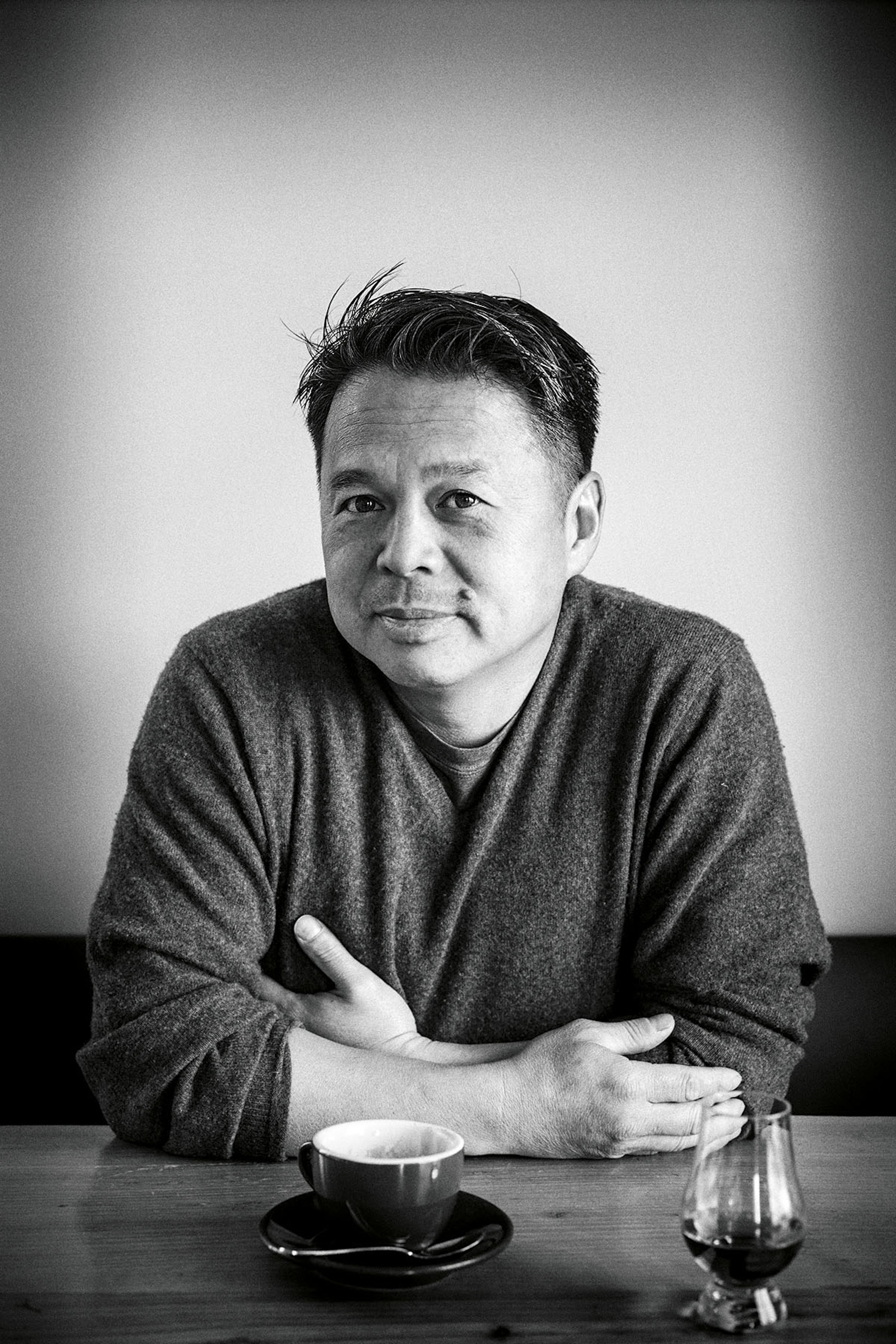 A black and white image of a person sitting at a table with a cup and glass. They are wearing a sweater and have arms crossed, looking at the camera.