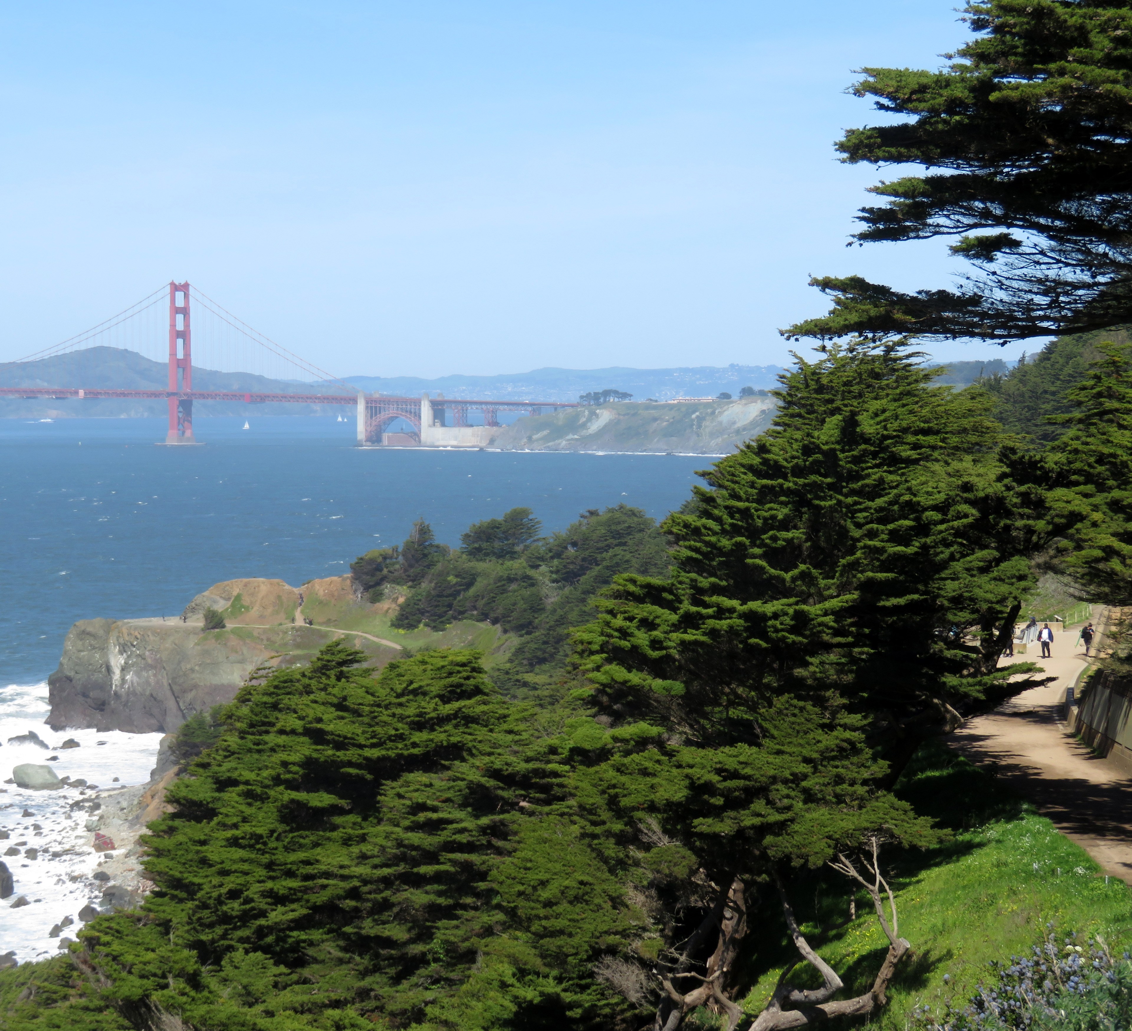 A coastal view shows a winding path bordered by lush trees, with the Golden Gate Bridge in the distance under a clear blue sky.