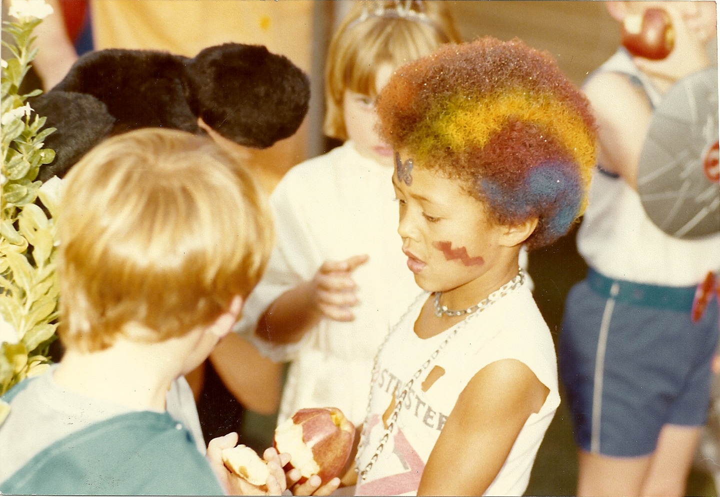 A group of children are gathered, one with a colorful afro and face paint talks to another with light hair. They're holding fruit in a lively outdoor setting.