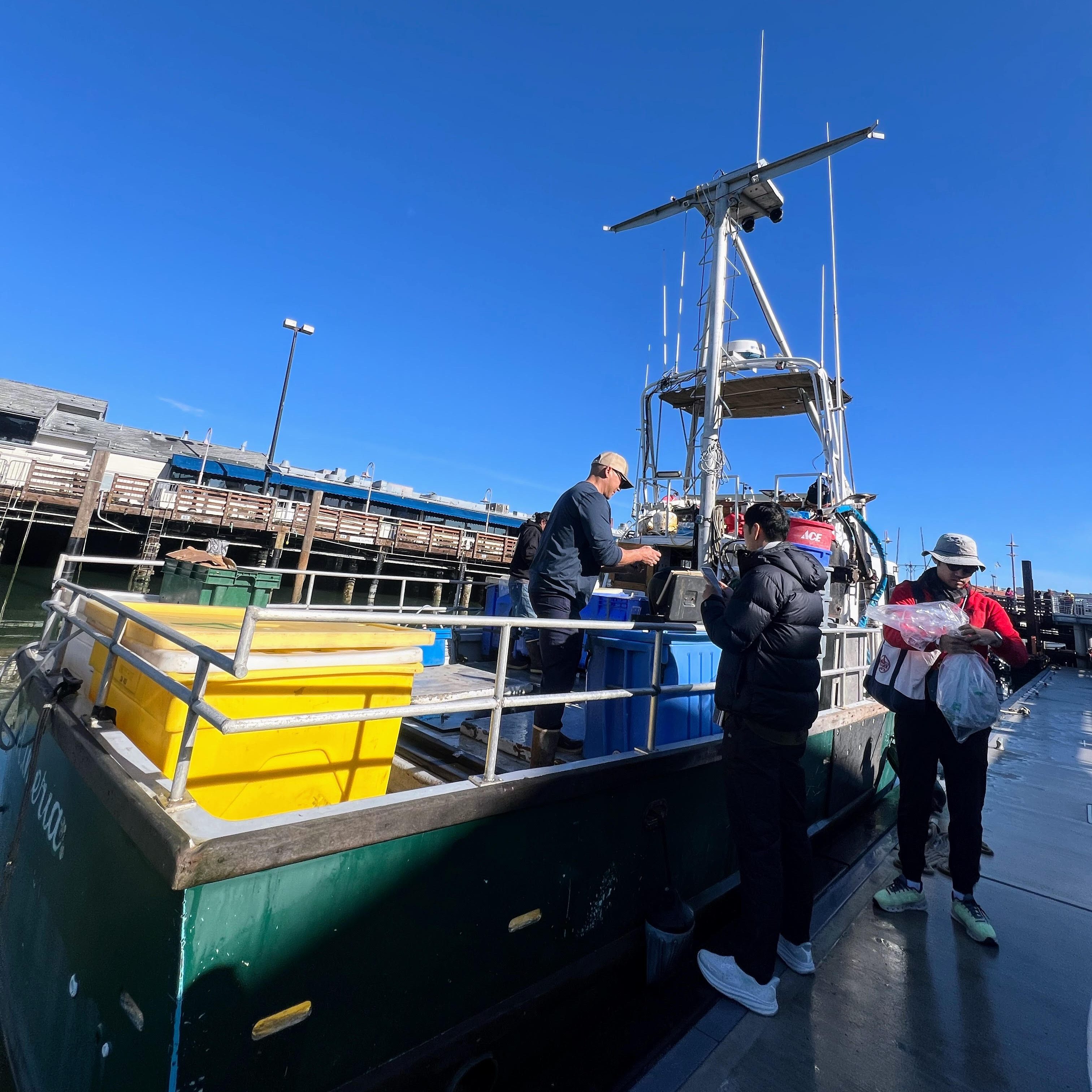 People stand on a dock next to a green boat filled with colored containers. One person is on the boat, and it's a clear, sunny day.