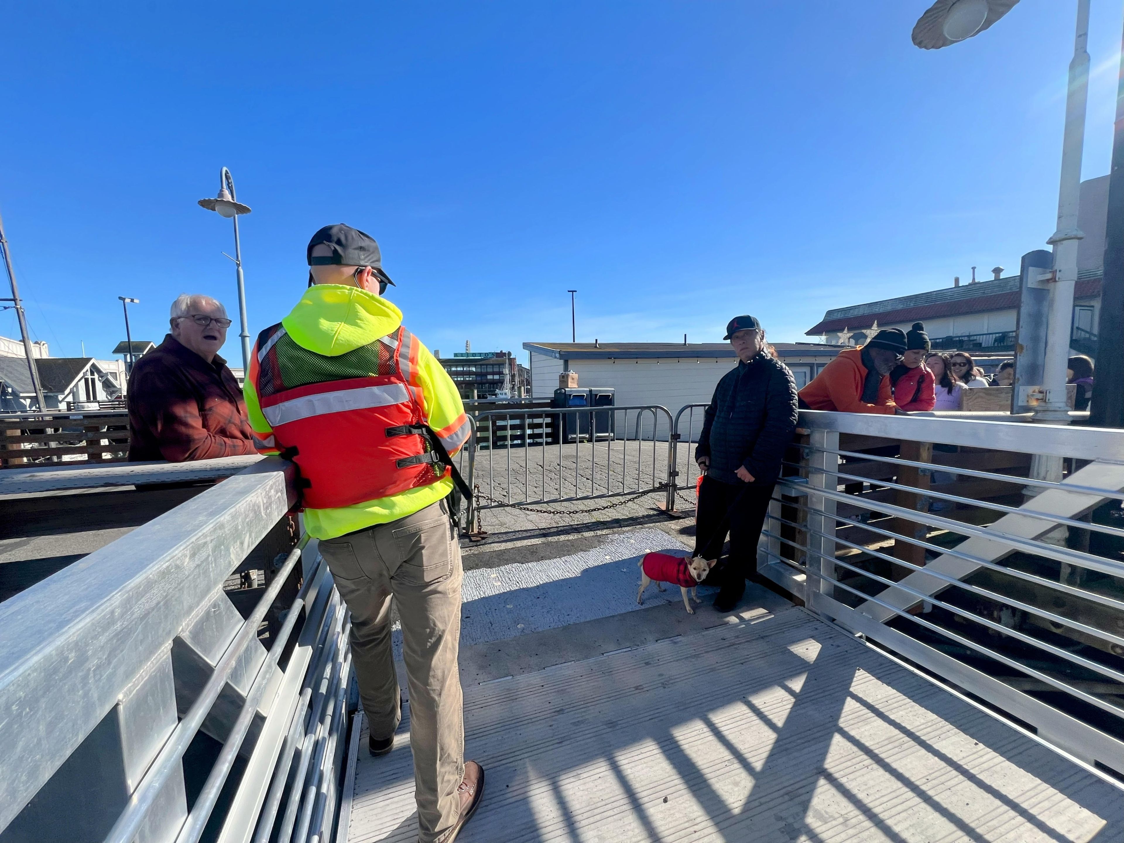 People are gathered on a sunny dock; one wears a high-visibility vest, others in casual attire, and a small dog in a red jacket stands by the railing.