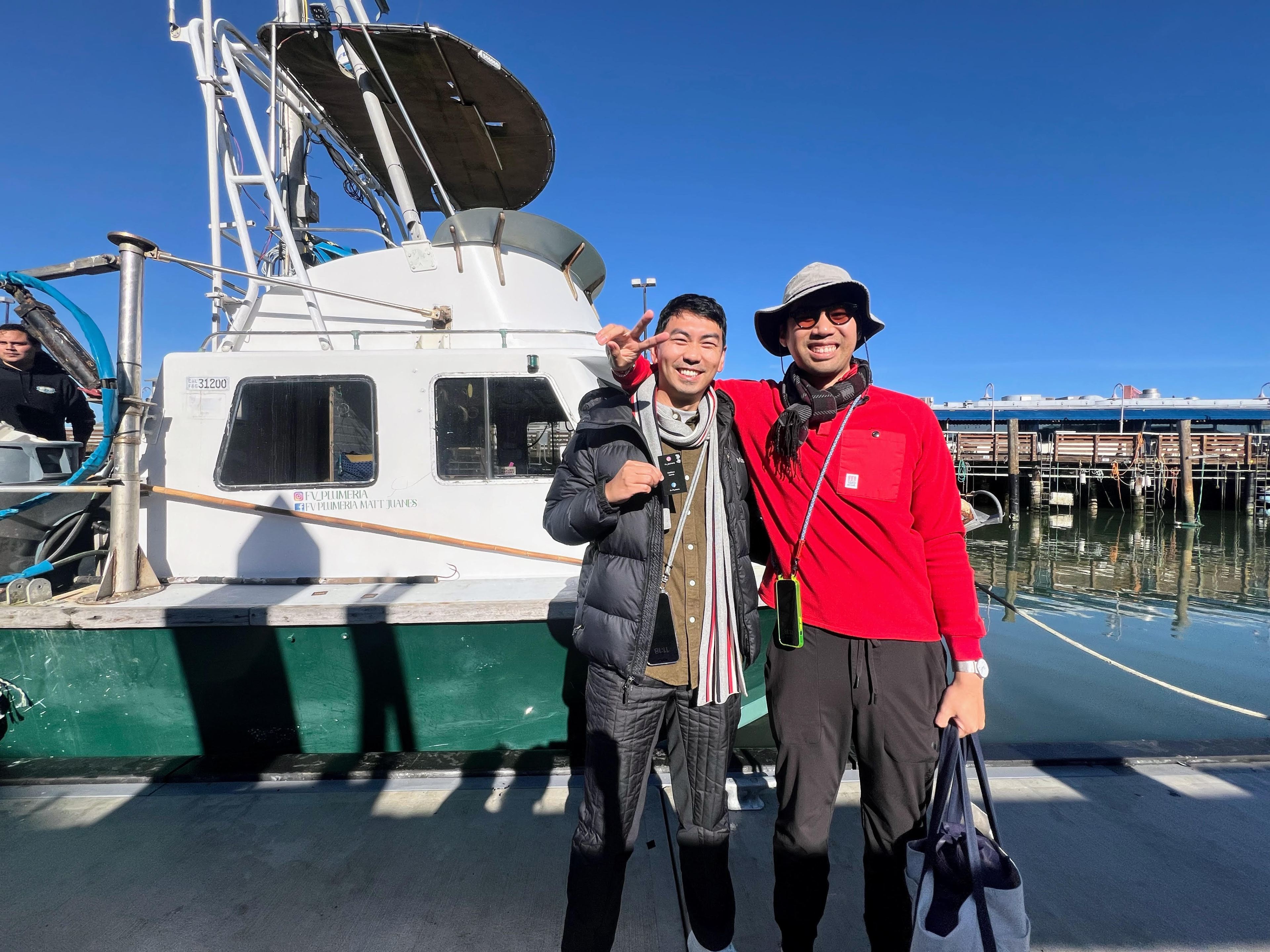 Two smiling individuals pose on a sunny dock, with a white boat behind them. One wears a black jacket, the other a red shirt and hat. The water is calm.