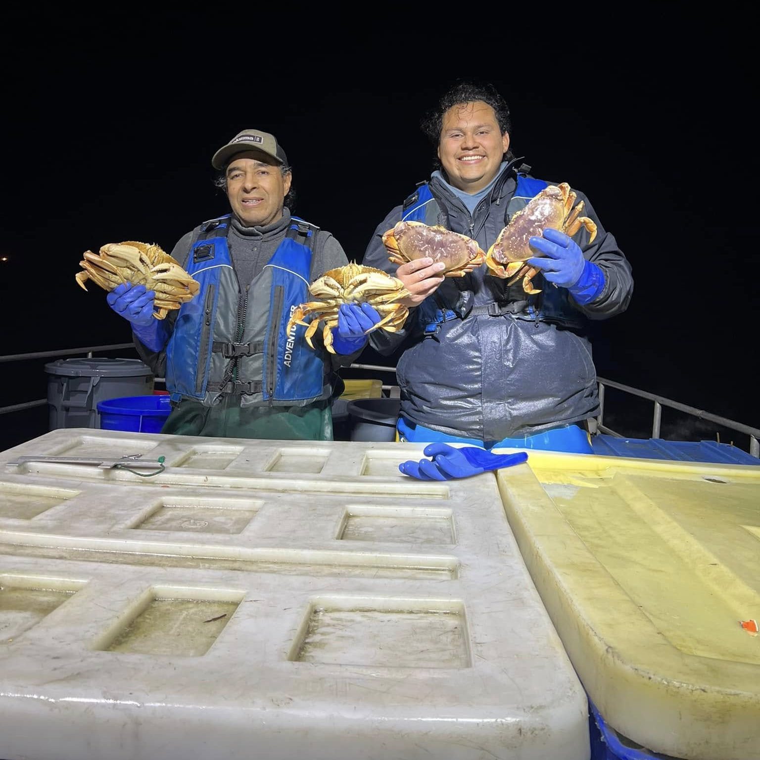 Two people wearing blue gloves and jackets are on a boat at night, smiling and holding several crabs each. There's a large cooler in front of them.