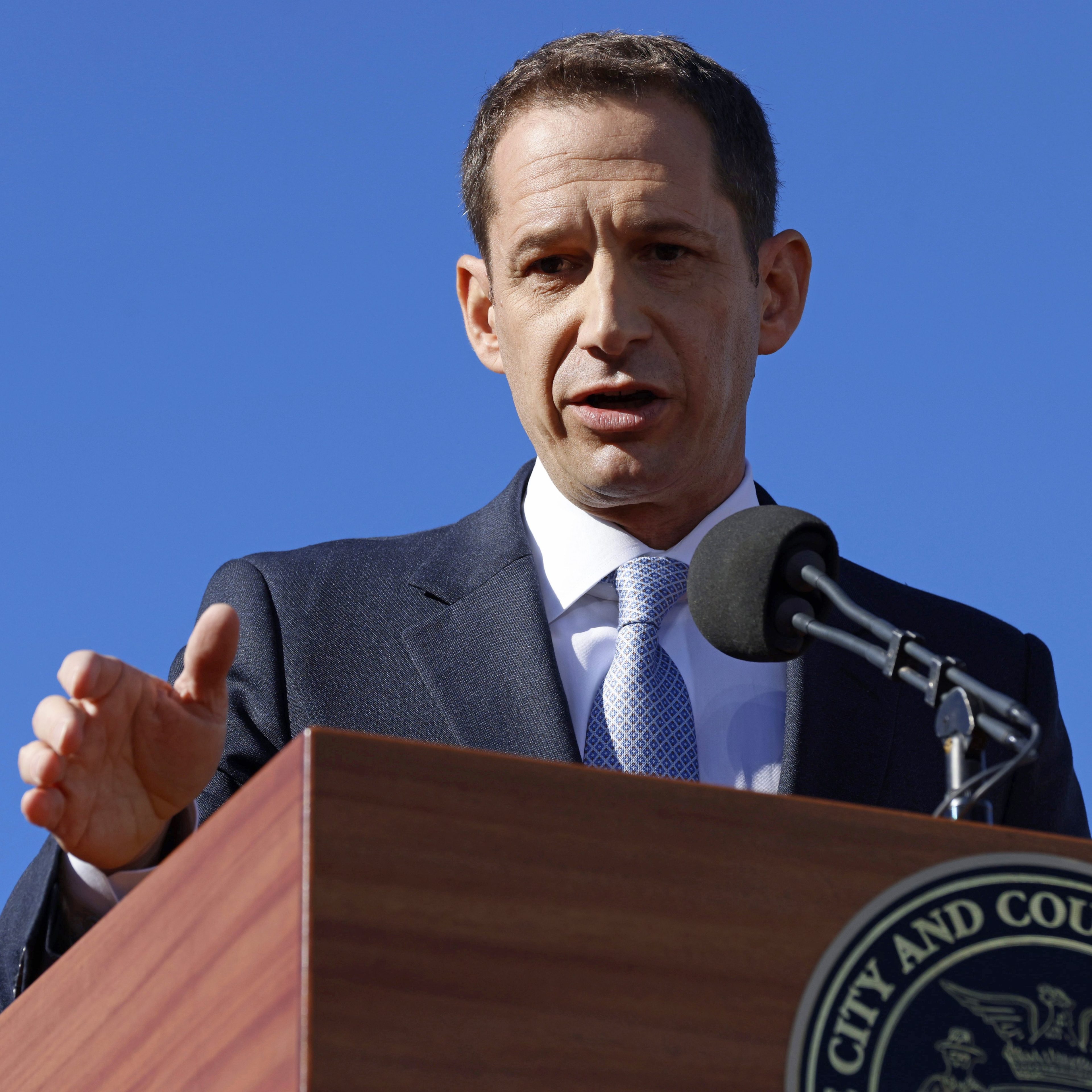 A person in a suit speaks at a podium with a microphone in front of a clear blue sky. The podium has an insignia for the "City and County of" an unspecified place.