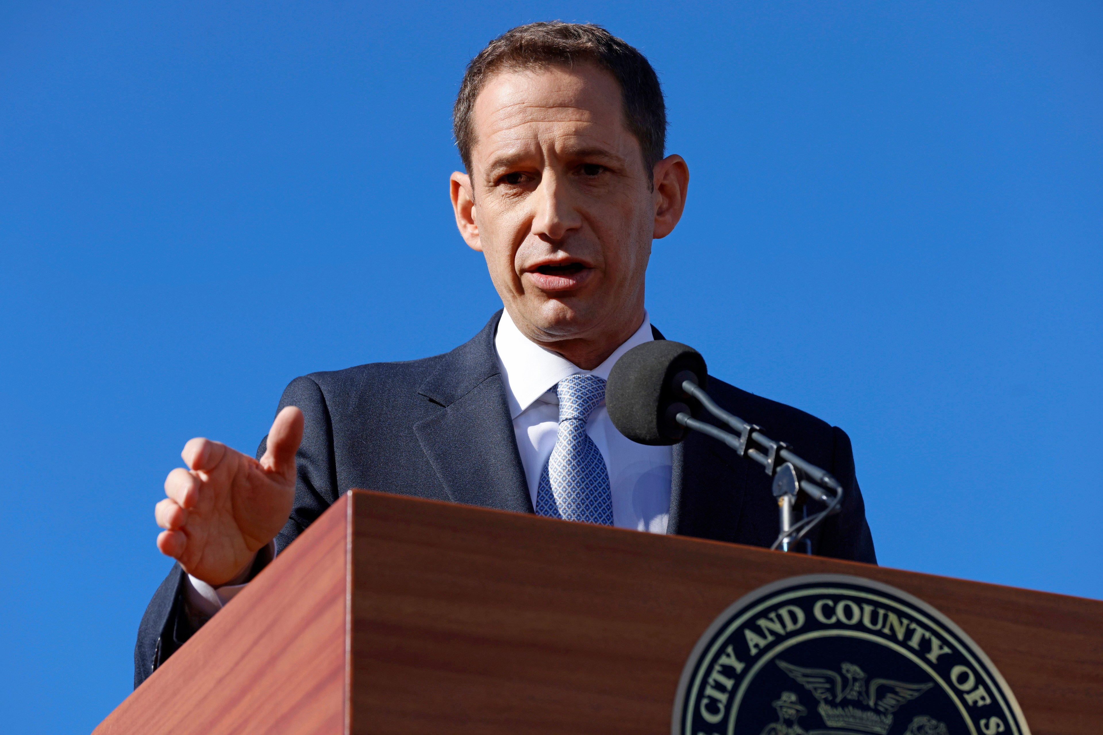 A person in a suit speaks at a podium with a microphone in front of a clear blue sky. The podium has an insignia for the &quot;City and County of&quot; an unspecified place.