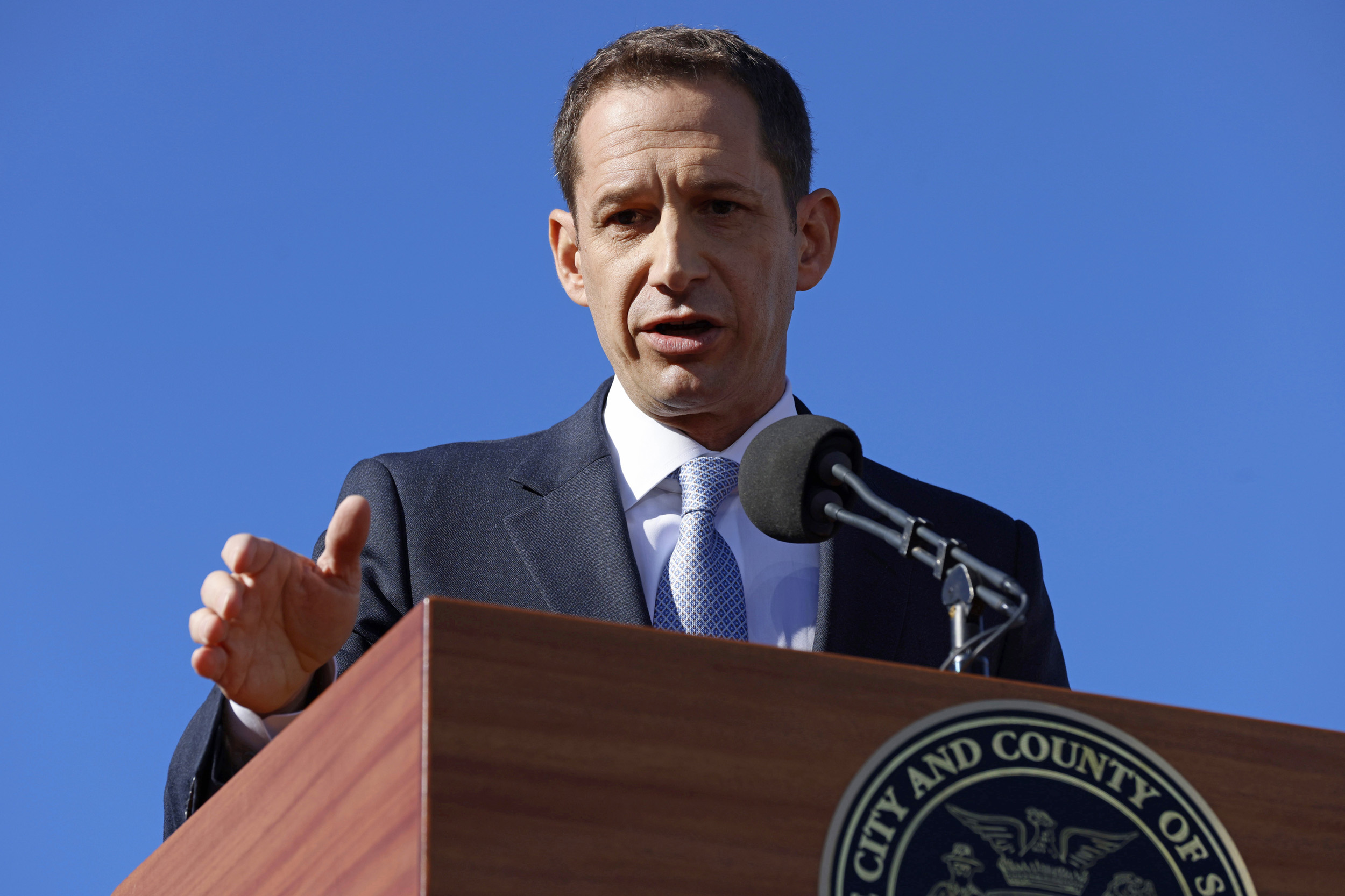 A man in a suit speaks at a podium with a microphone against a clear blue sky. The podium bears the seal of "City and County of SF."