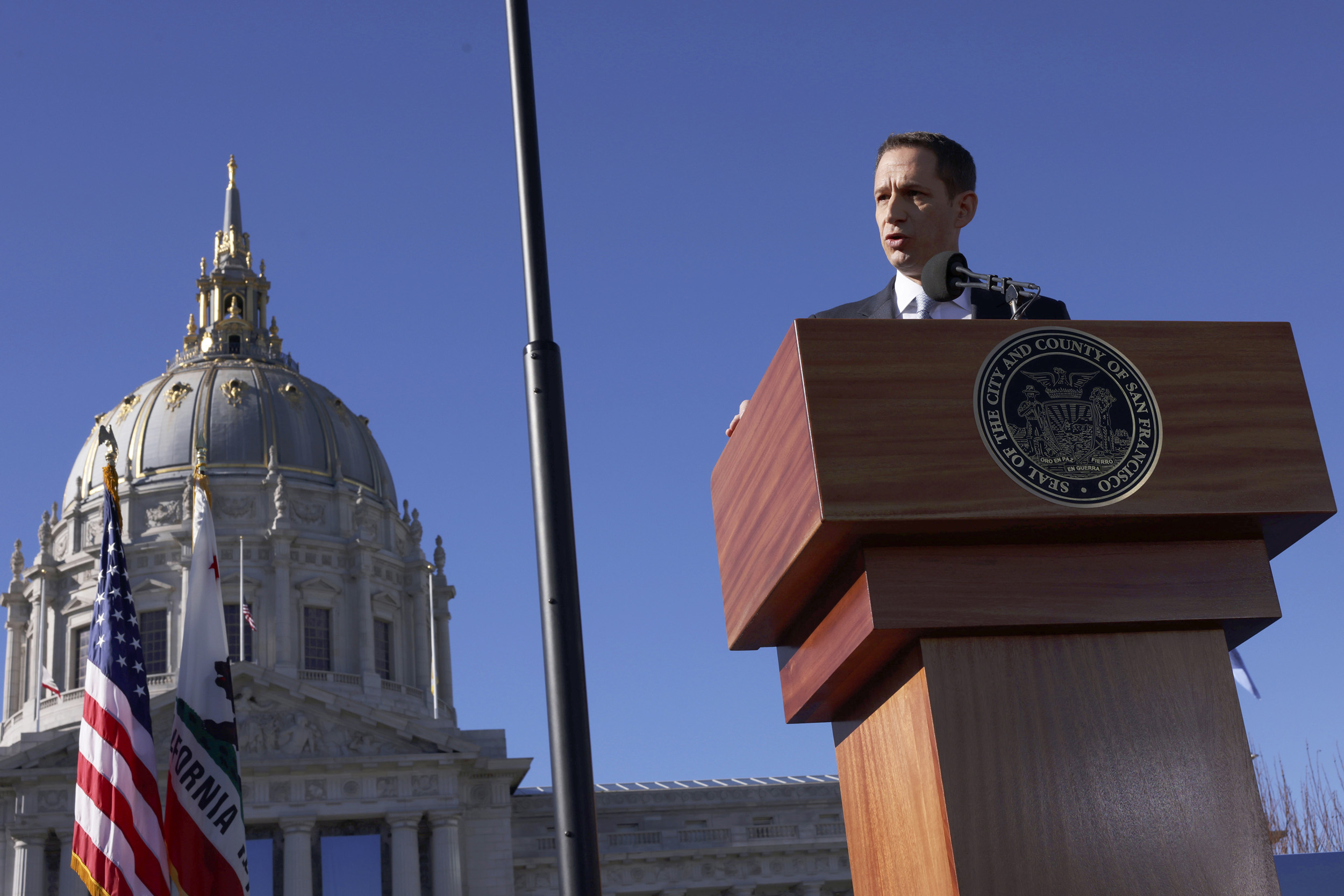 A man is seen from the shoulders up standing behind a lectern with San Francisco City Hall in the background.