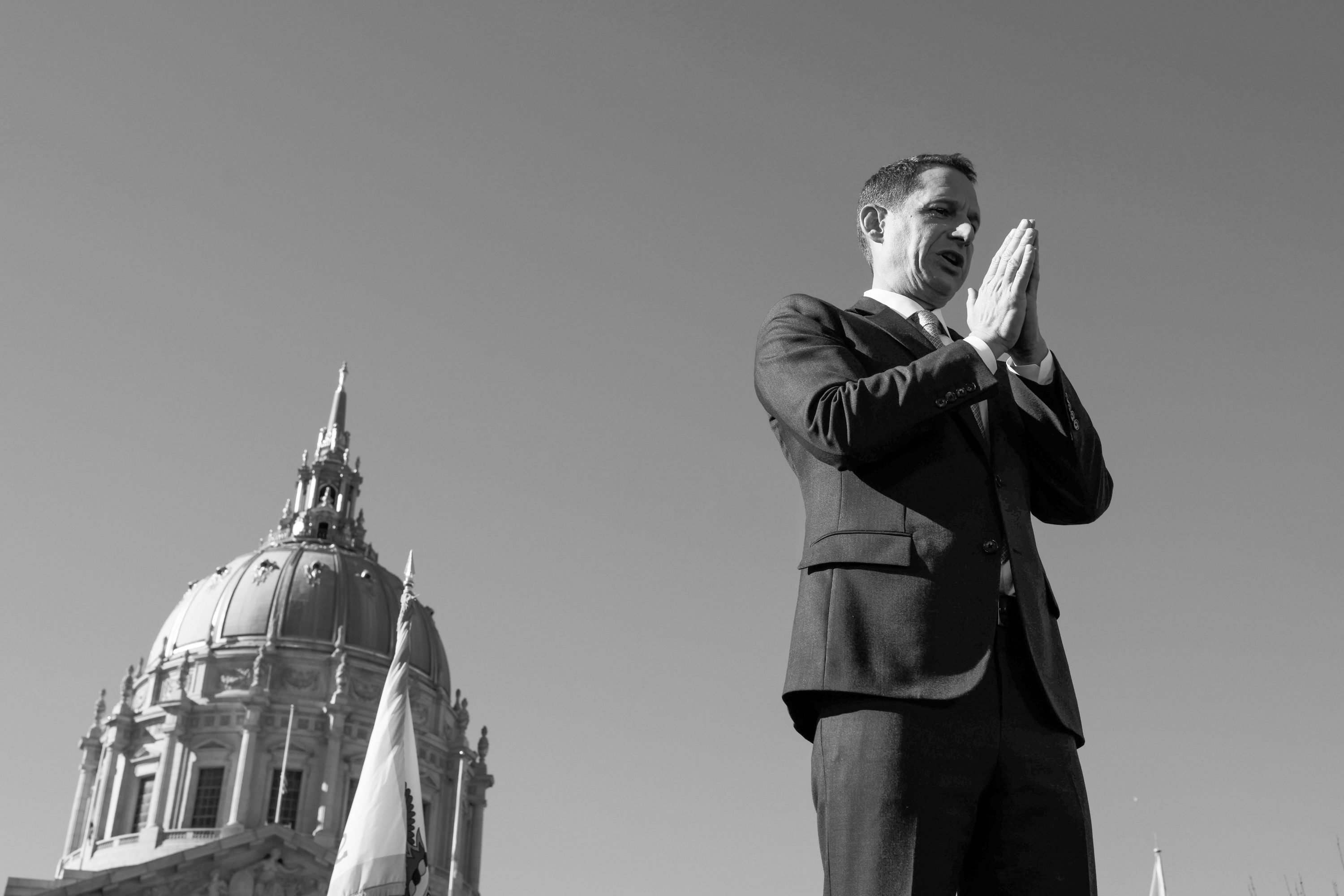 A man in a suit stands with hands clasped near his face, speaking or presenting, with a large domed building and flags in the background under a clear sky.