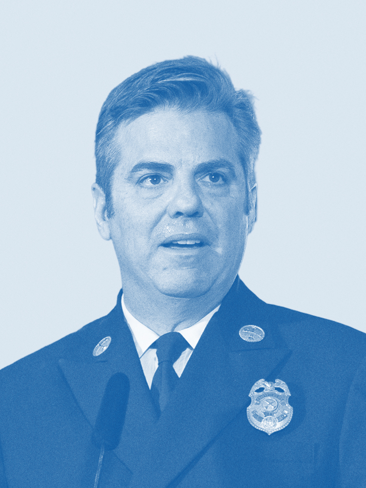 A person in a fire department dress uniform is speaking, with a microphone visible. The image has a blue tint.