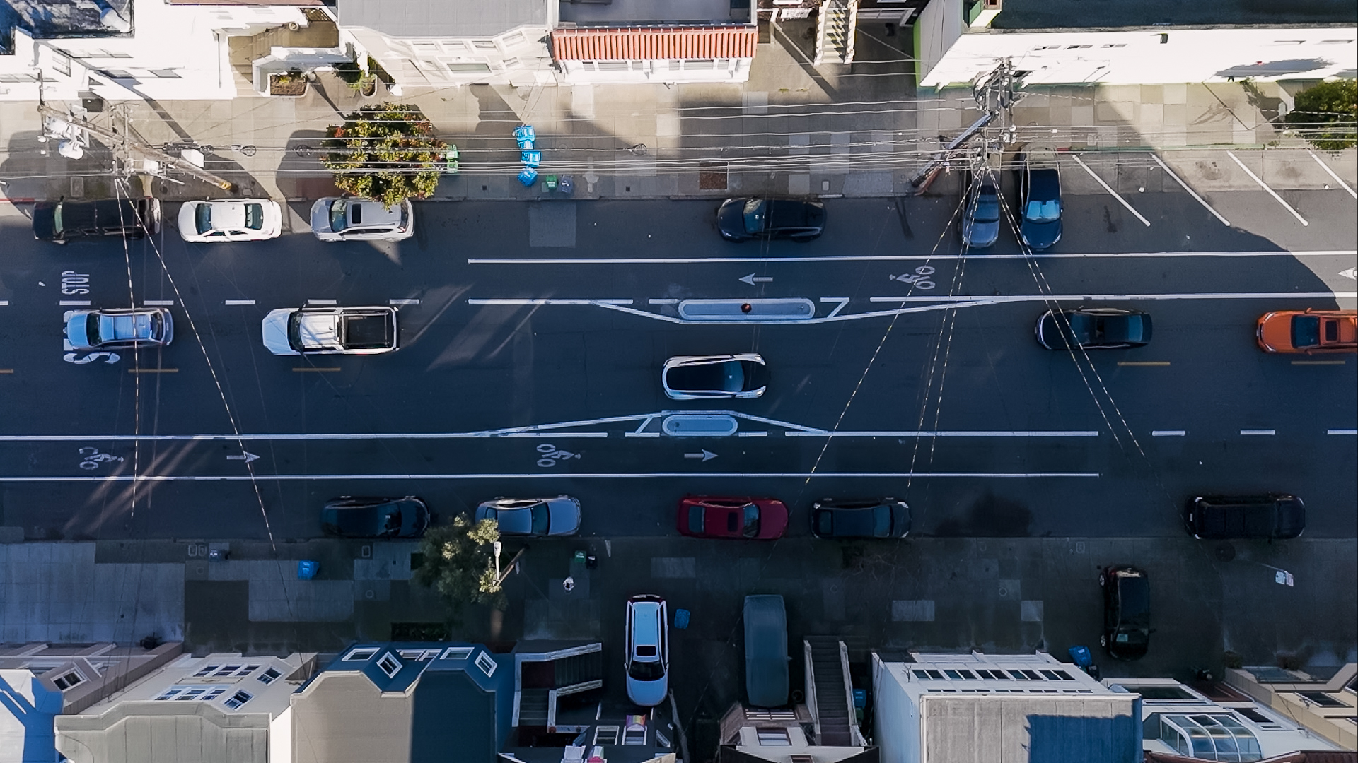 This is an aerial view of a street with parked cars on both sides, moving traffic in the center, and visible bike lanes. Buildings line the sidewalks.