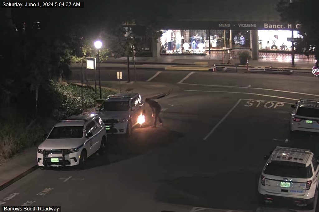 A nighttime street scene shows a person using a tool that emits sparks near a vehicle, with police cars parked nearby and a closed lane sign in view.