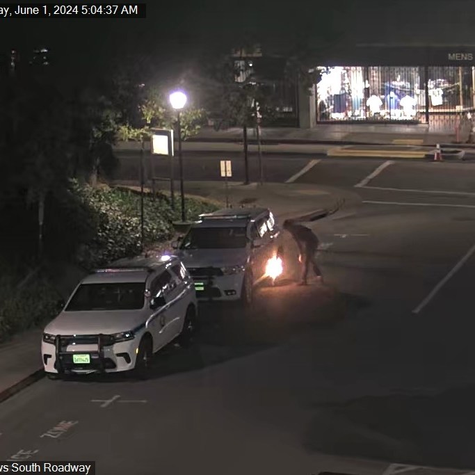 A nighttime street scene shows a person using a tool that emits sparks near a vehicle, with police cars parked nearby and a closed lane sign in view.