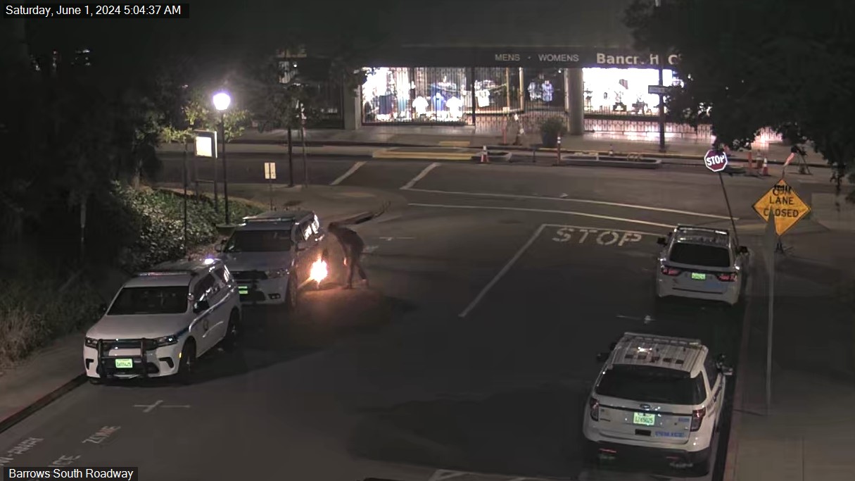 A nighttime street scene shows a person using a tool that emits sparks near a vehicle, with police cars parked nearby and a closed lane sign in view.