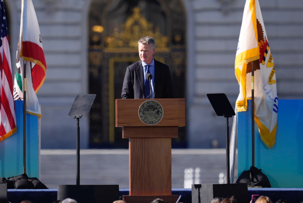 A person stands at a podium outdoors, flanked by two flags. They are speaking into a microphone, with a formal building behind them.