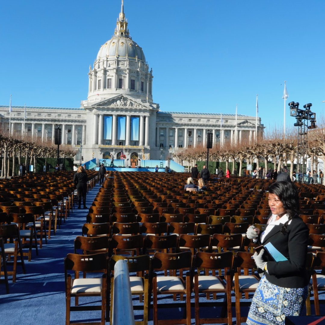 Rows of wooden chairs face a grand, domed building under a clear blue sky. A few people walk among the chairs on a blue carpet; a woman in foreground.