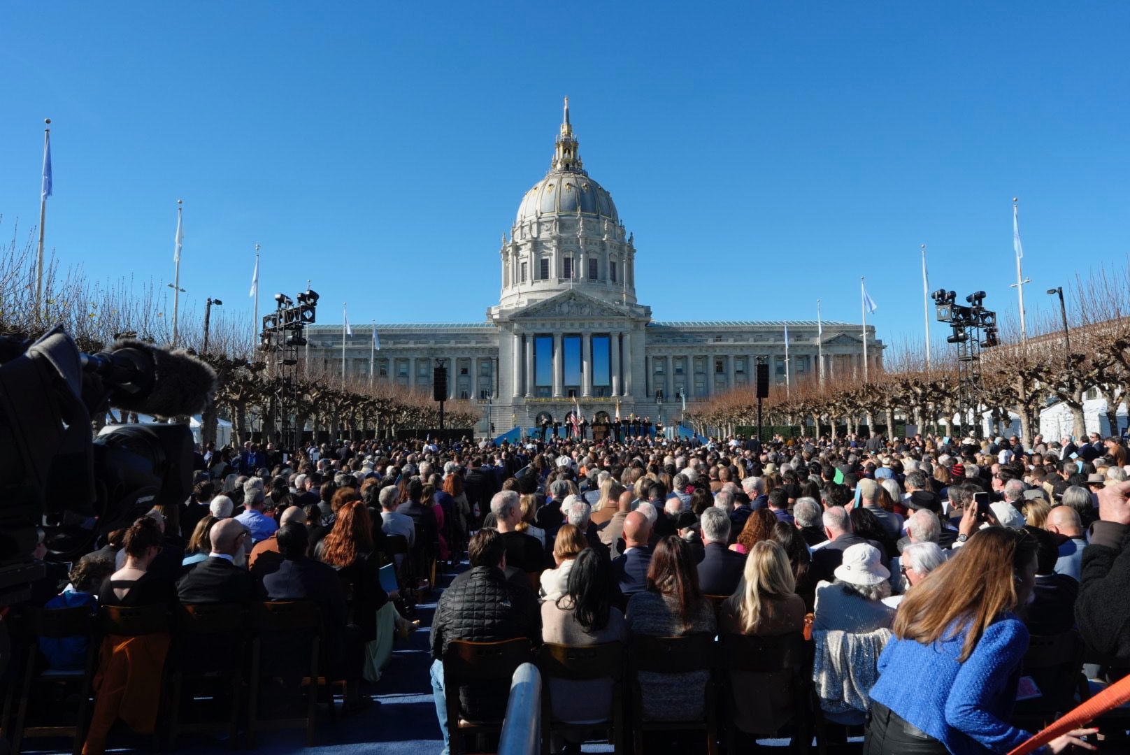 A large crowd sits facing a grand building with a dome under a clear blue sky. Flags line the pathway, and a camera is visible on the left side.