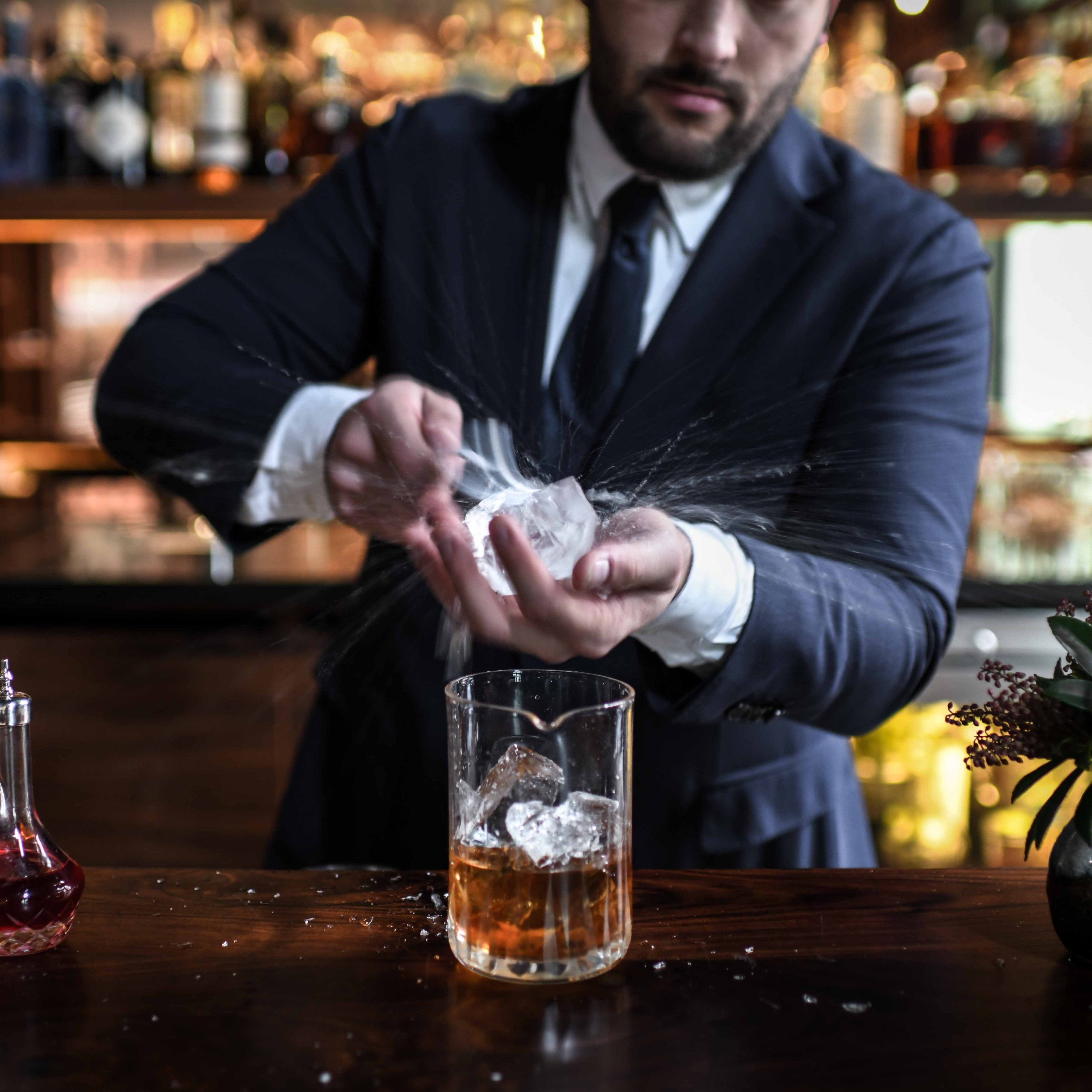 A bartender in a suit carves a large ice cube, creating splashes. Glass decanters with colored liquids and a citrus-filled vase adorn the bar counter.