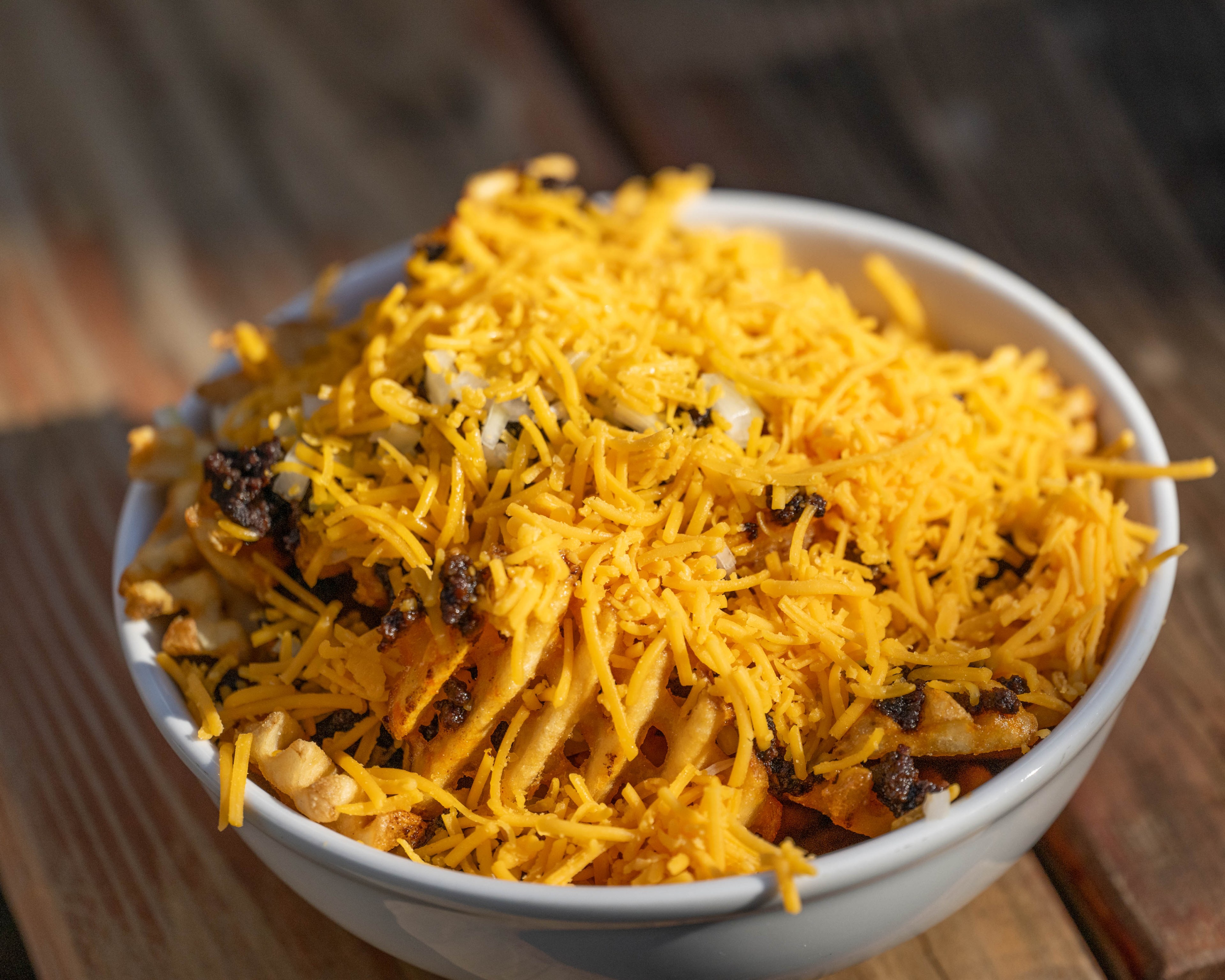 A bowl of waffle fries is covered with a generous amount of shredded cheese and topped with bits of ground meat, sitting on a wooden surface.