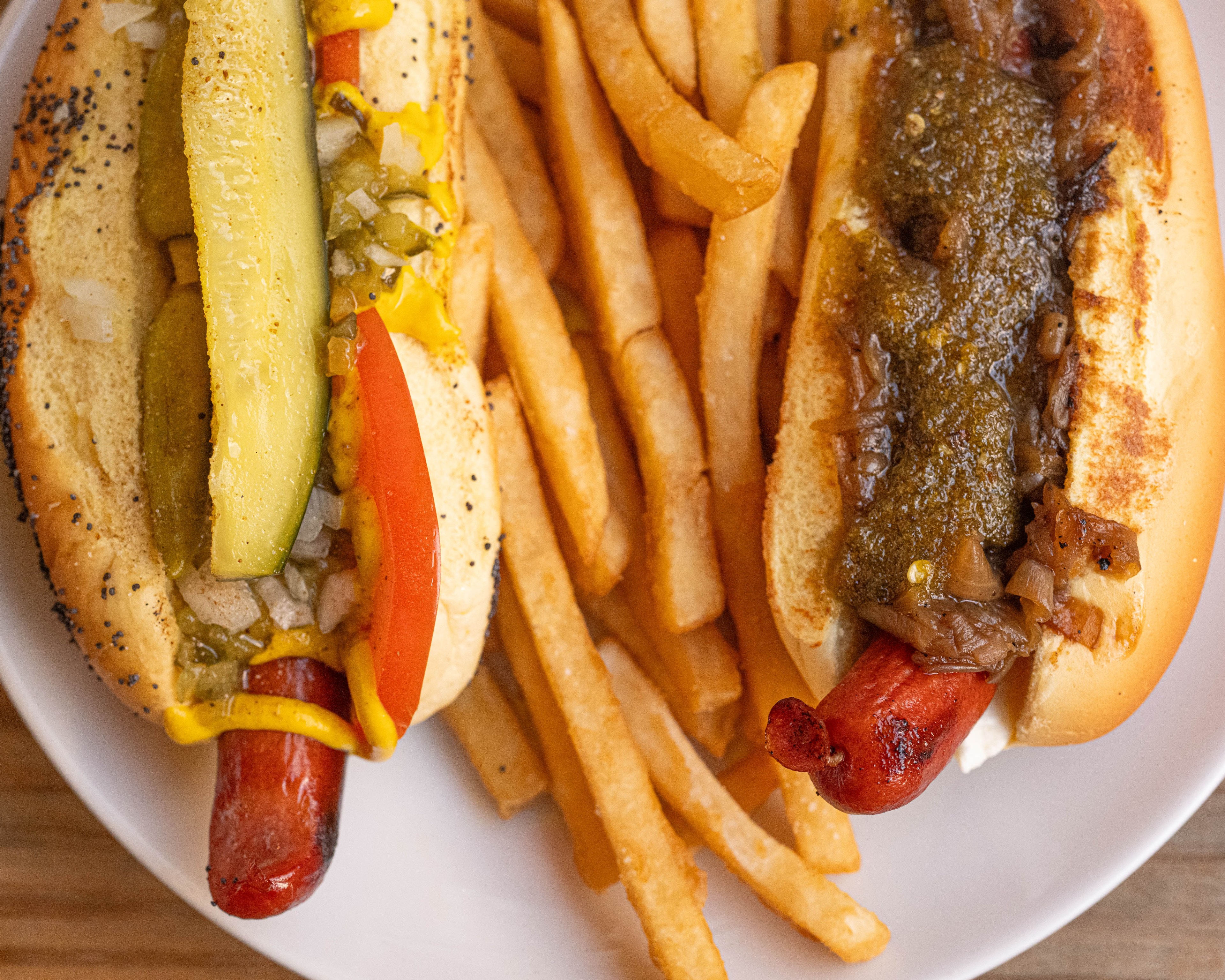 Two hot dogs with toppings, one with pickle, mustard, and tomato, the other with relish and onions, served with fries on a white plate.