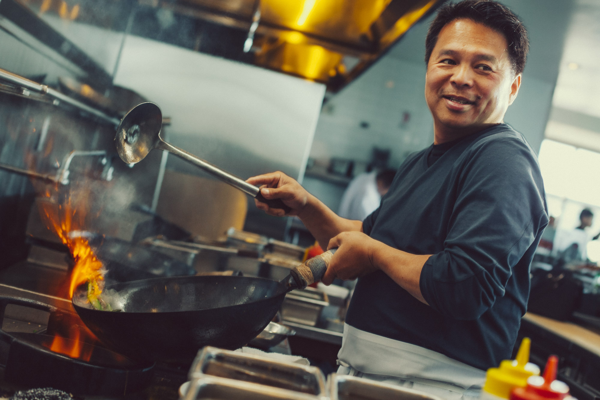 A chef smiles while stir-frying in a wok over a flaming stove in a commercial kitchen. He holds a ladle, and condiment bottles are in the foreground.