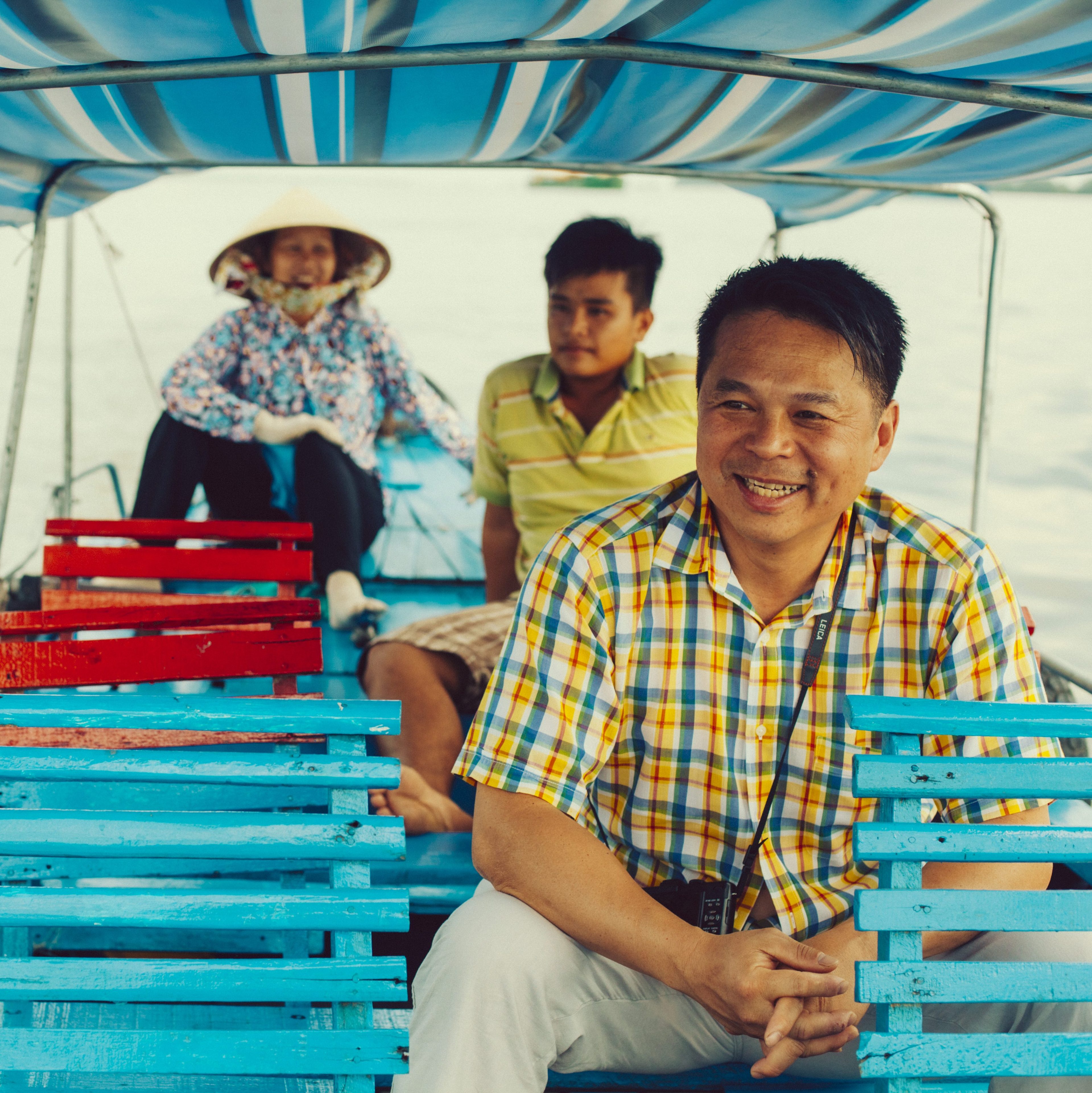 Three people sit on a boat under a striped canopy. The front man wears a colorful checkered shirt, smiling, with blue benches and two others behind him.