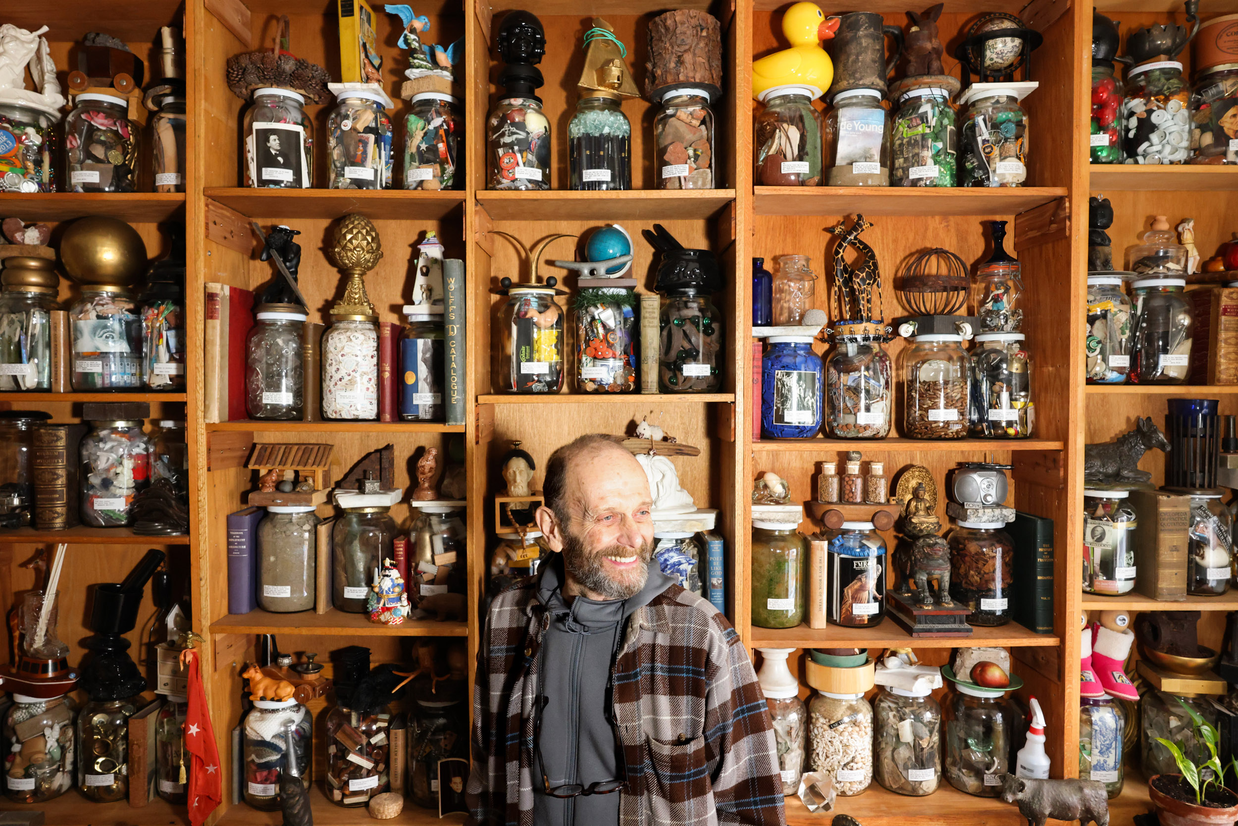 A man smiles in front of wooden shelves filled with various jars, each containing eclectic items like toys, artifacts, and decorative objects.