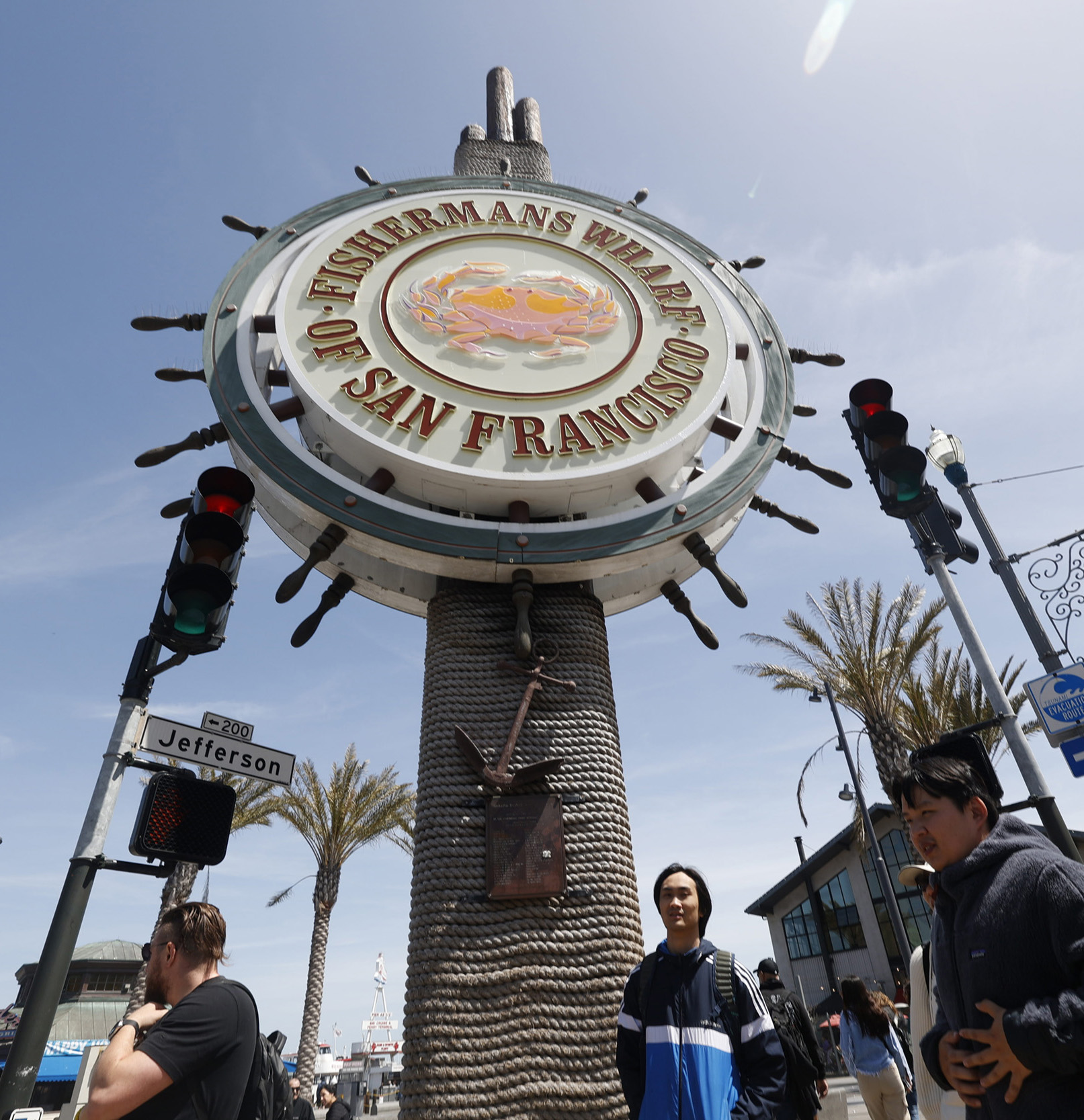 The image shows the Fisherman's Wharf sign in San Francisco, resembling a ship's wheel. Below, there's a street sign for Jefferson. People and palm trees are visible.