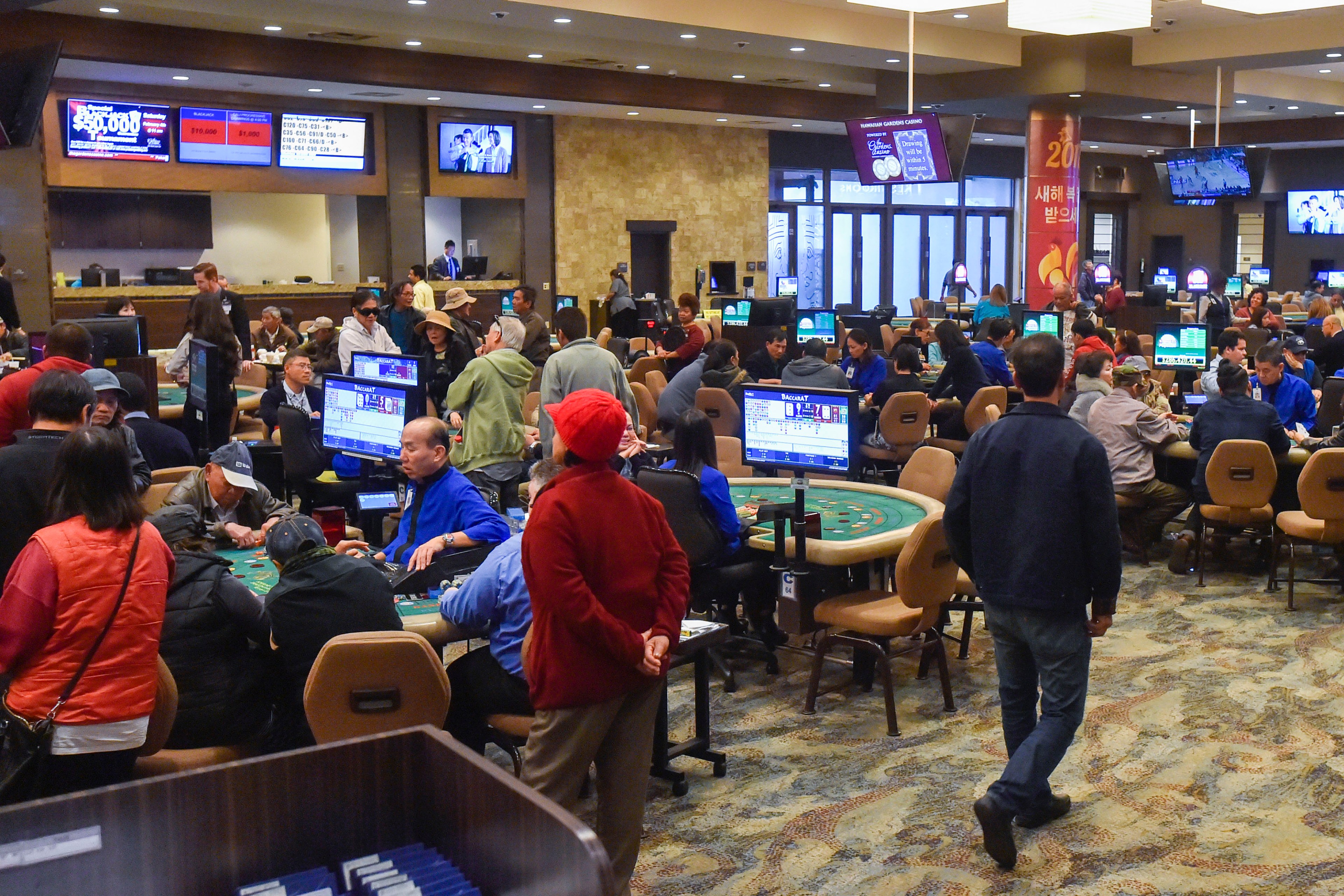 The image shows a busy casino with numerous people sitting and playing games at tables. There are large screens displaying information, and the atmosphere is lively.