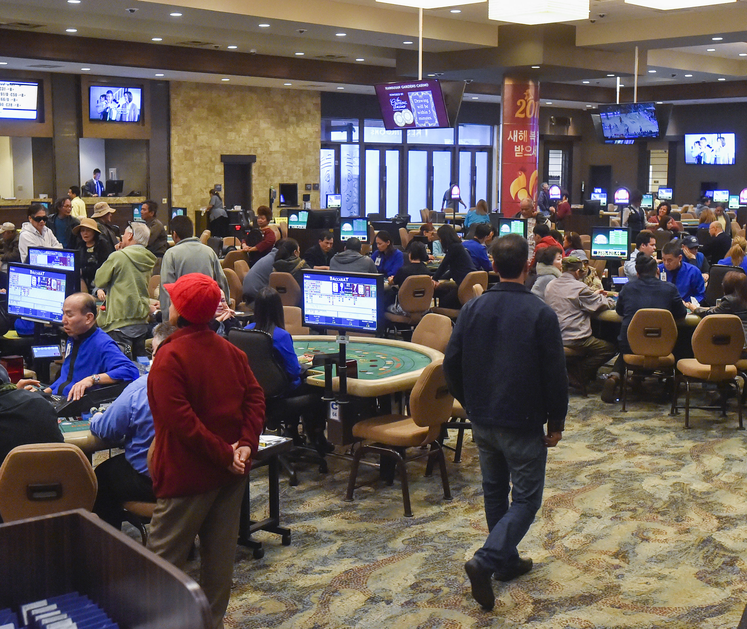 A busy casino floor with people seated at gaming tables and slot machines. Monitors display game stats, while others stand or walk through the area.