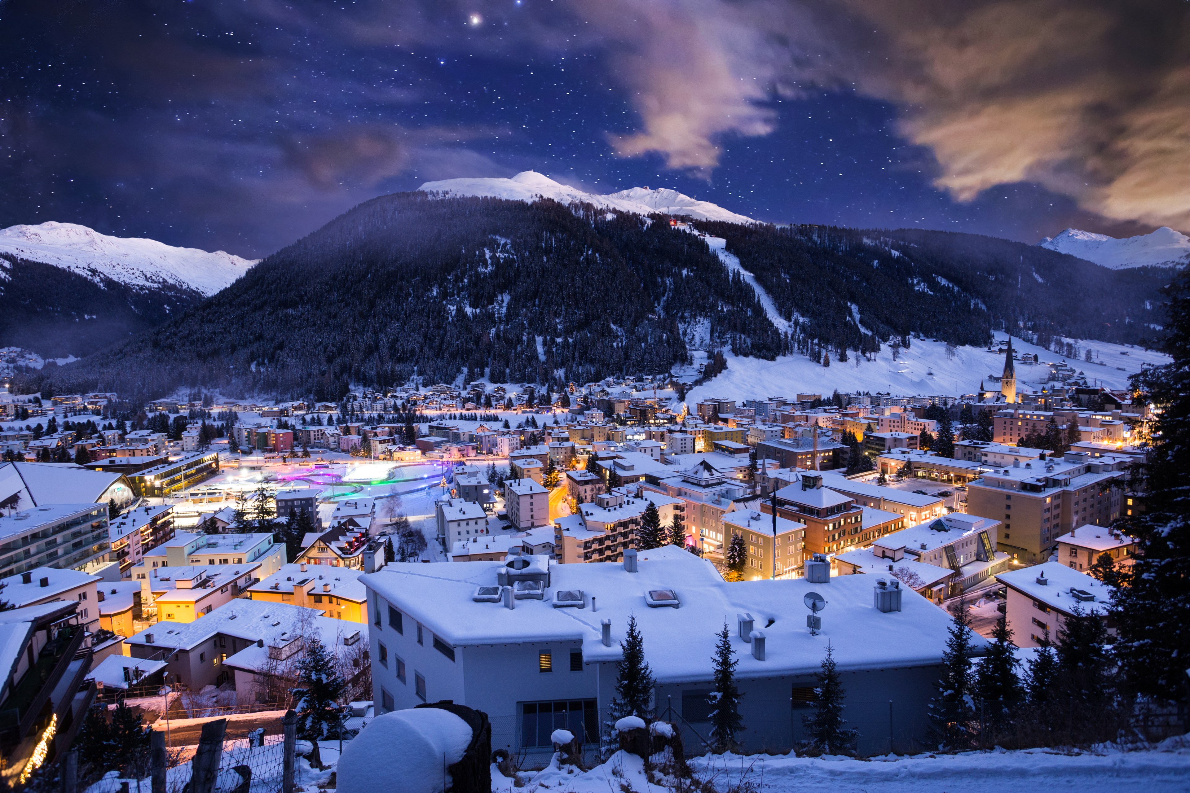 A snowy town is illuminated by colorful lights at night. Mountains rise in the background beneath a starry sky with a few clouds.