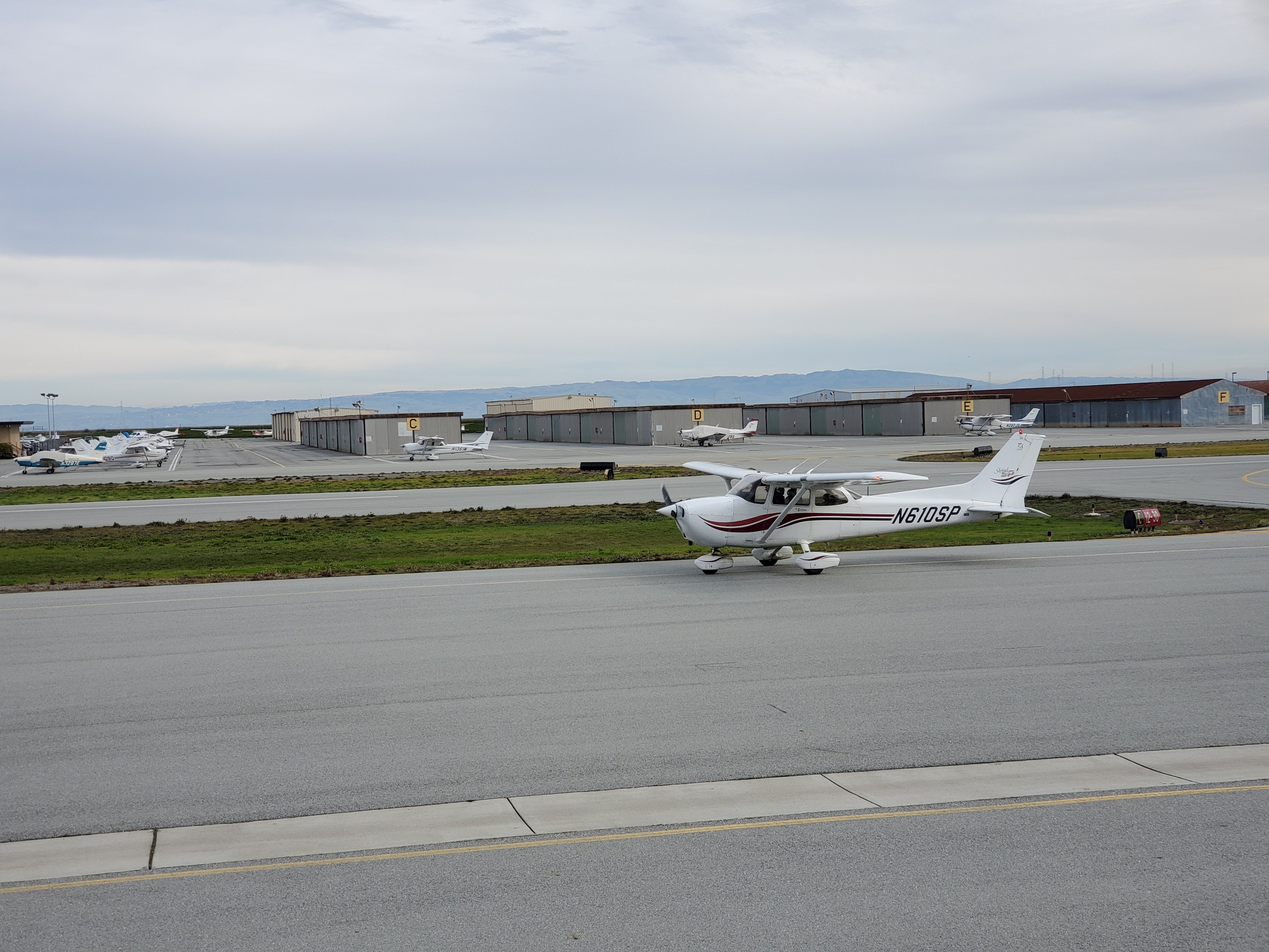 A small white airplane with red accents is on a taxiway at an airport, with hangars and parked planes in the background under a cloudy sky.