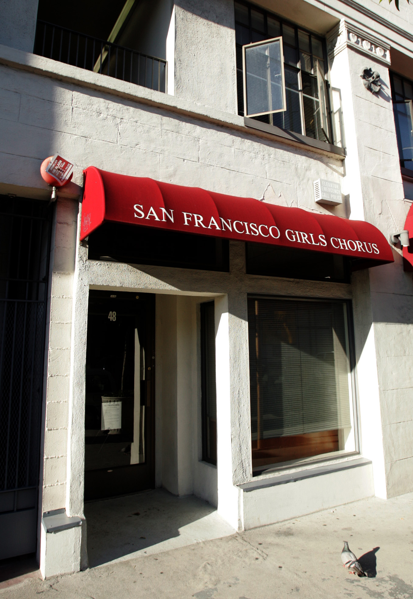 A building with a red awning reads "San Francisco Girls Chorus." There's a glass door, adjacent window, and a pigeon on the sidewalk.