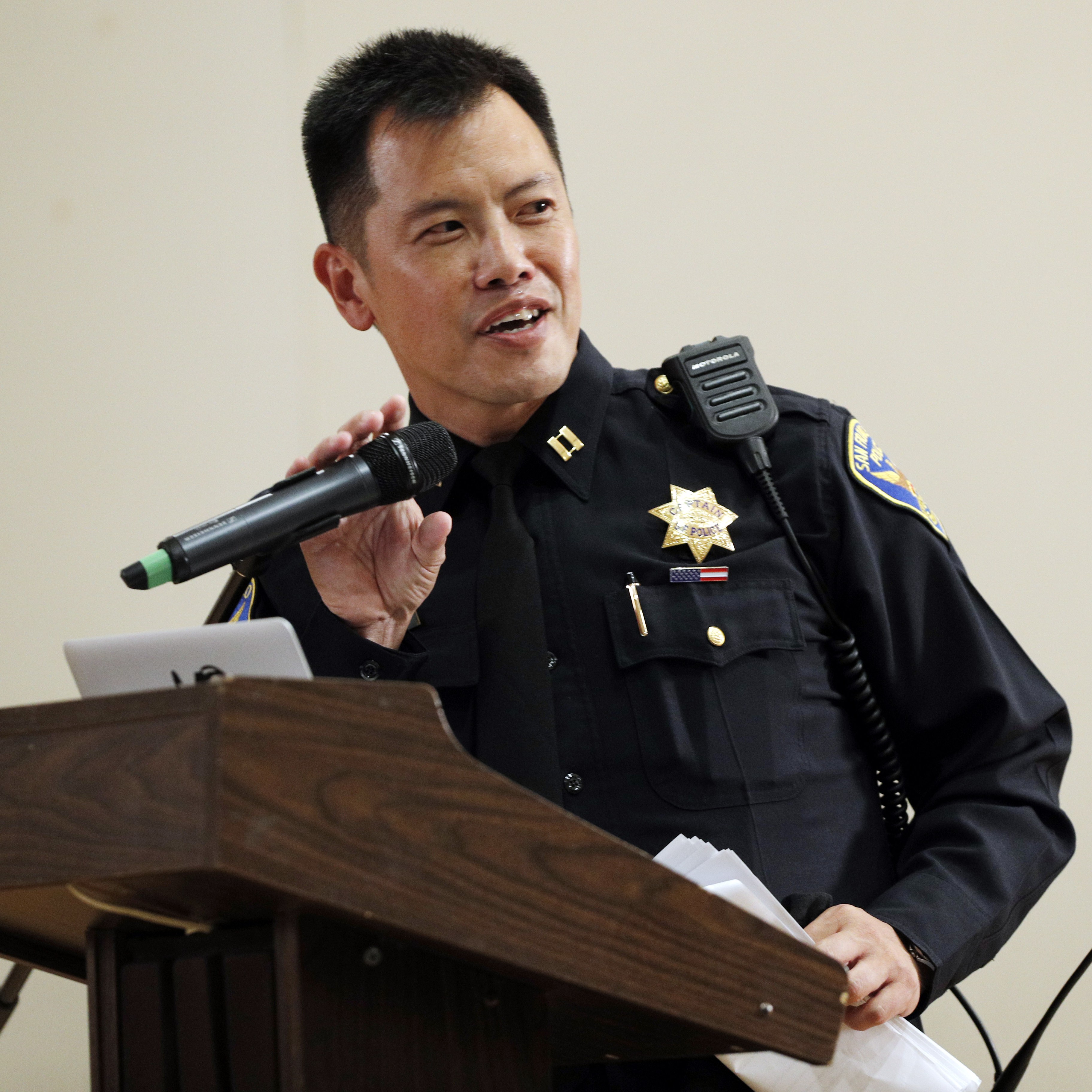 A police officer in uniform speaks into a microphone at a wooden podium, holding papers. He wears a badge and radio on his chest.