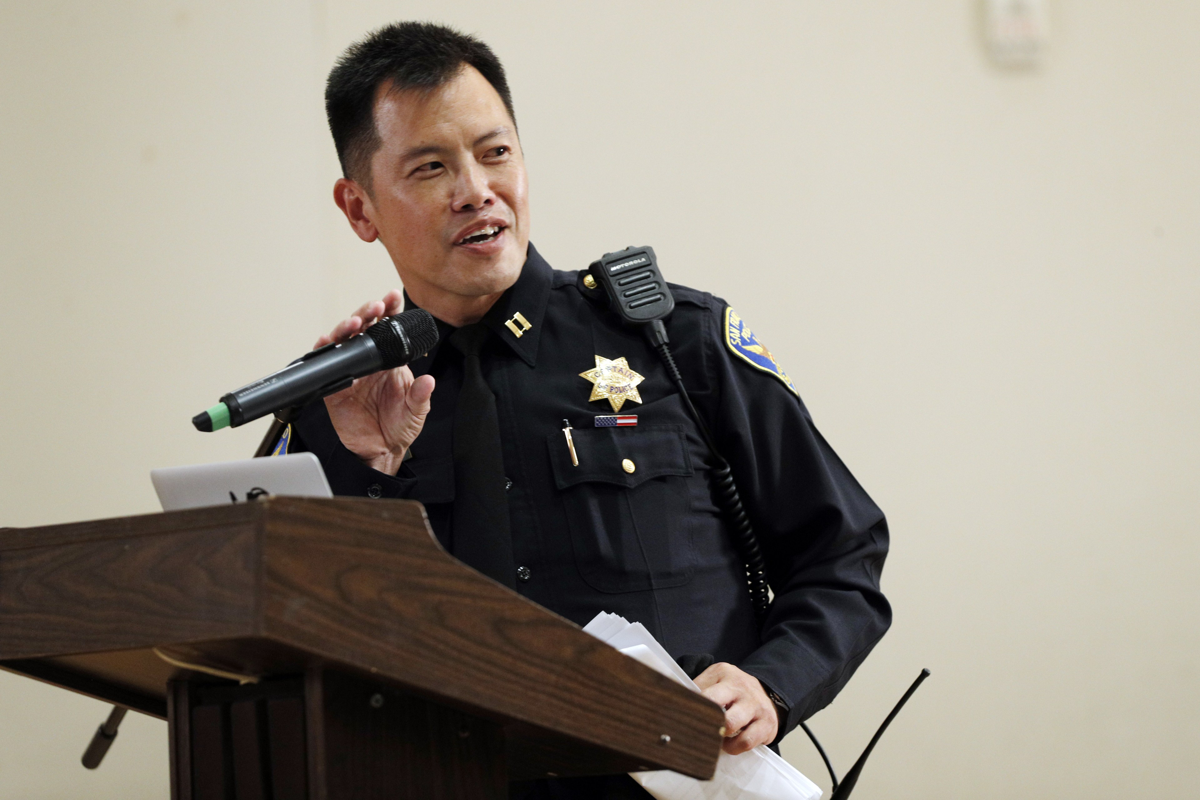 A police officer in uniform speaks into a microphone at a wooden podium, holding papers. He wears a badge and radio on his chest.