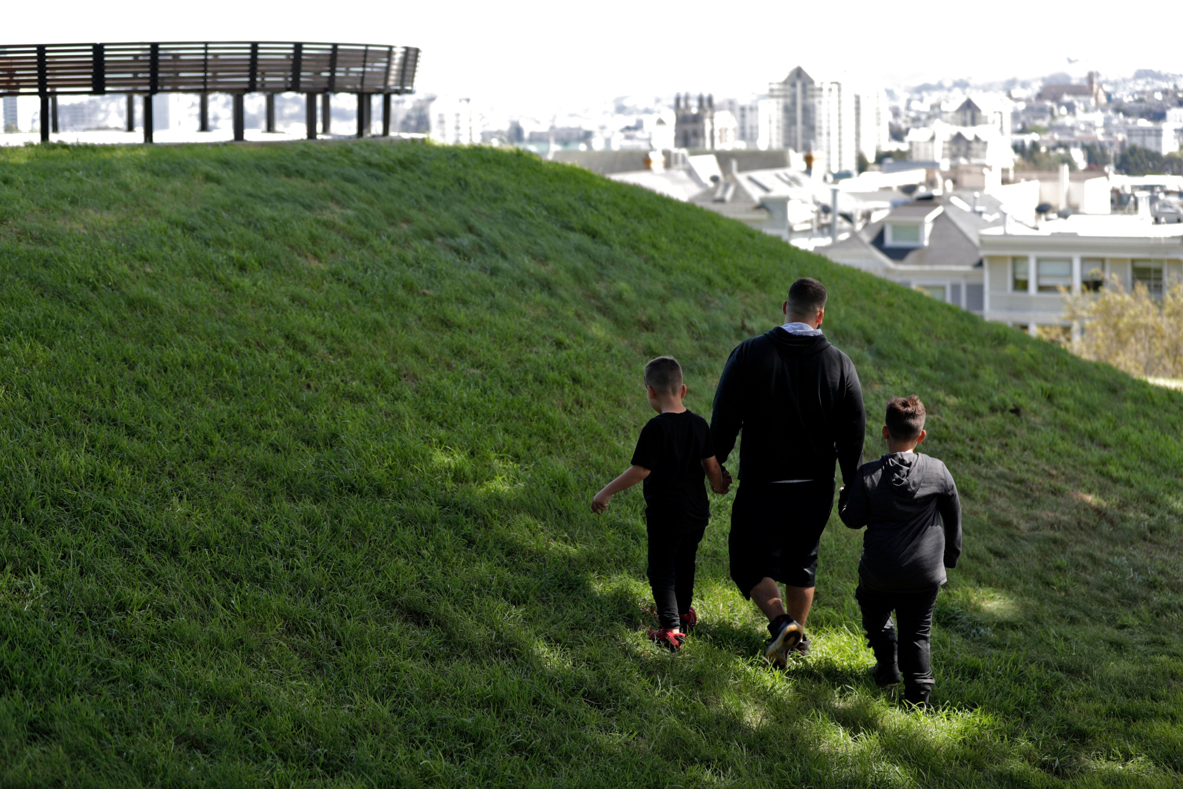 A man and two children walk hand in hand on a grassy slope, with city buildings visible in the background under a bright sky. A curved bench sits atop the slope.