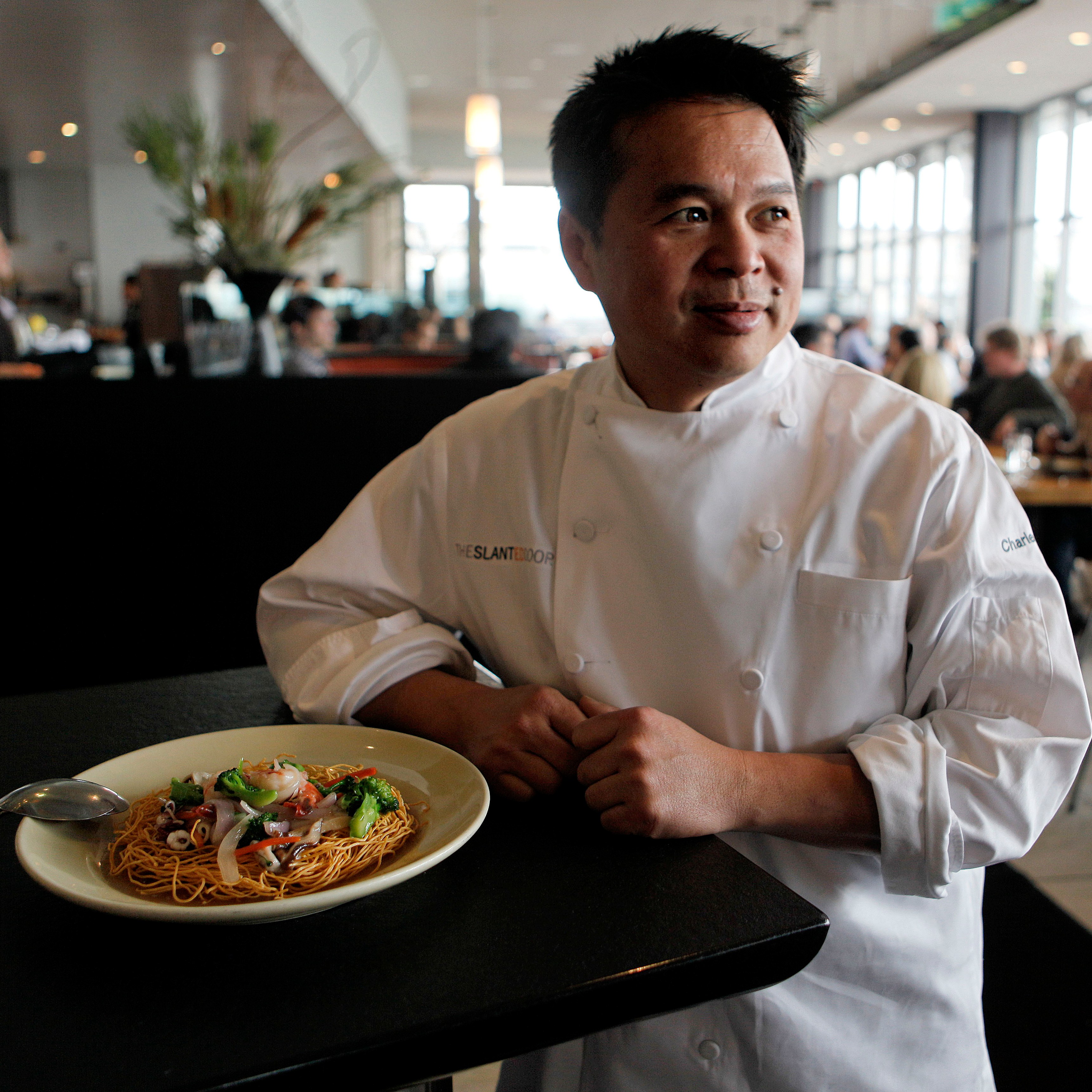 A chef in a white uniform stands beside a plate of noodles garnished with vegetables, in a busy restaurant with people dining in the background.