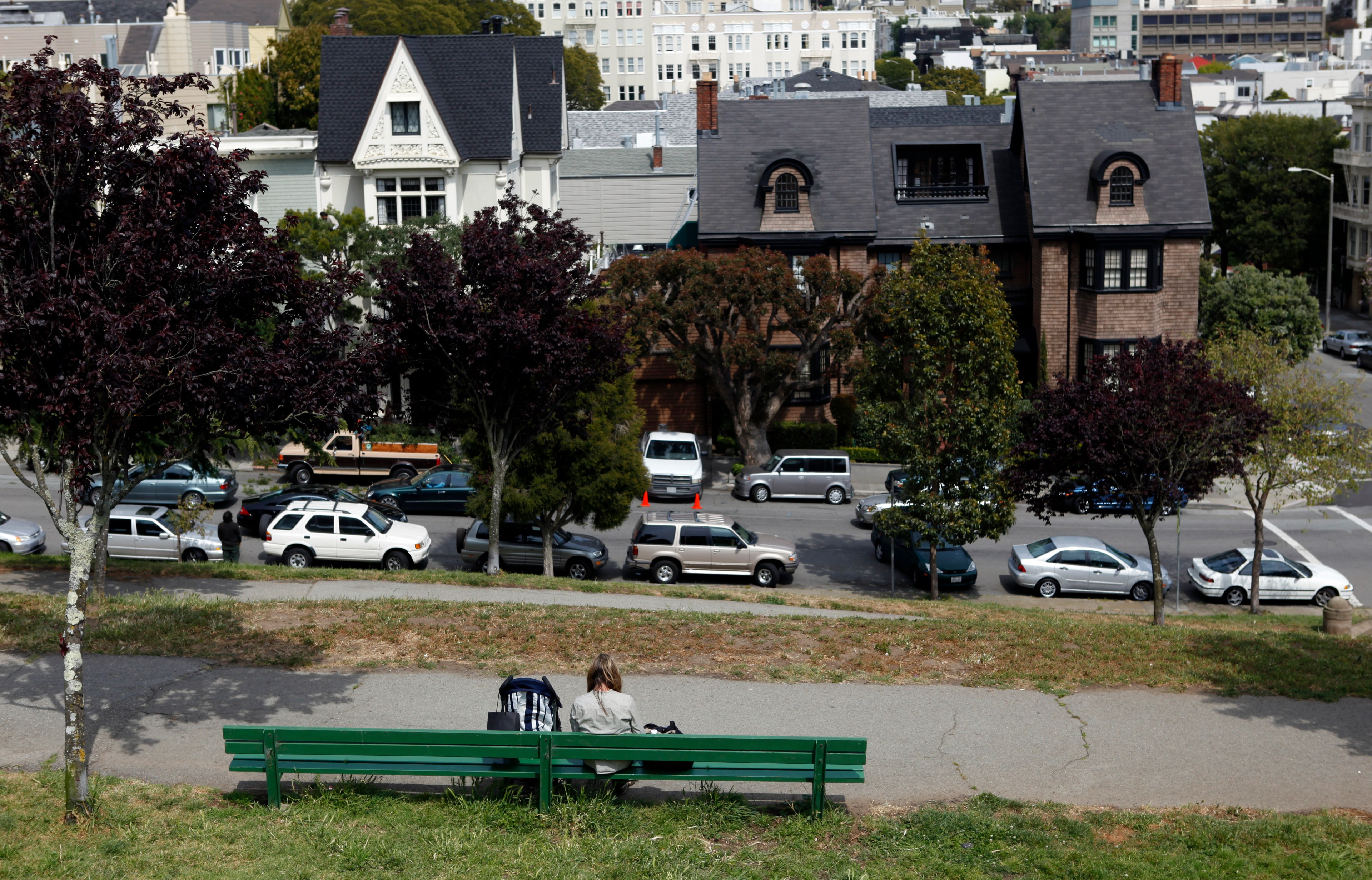 A person sits on a green bench in a park, facing a quiet street lined with parked cars. Two charming houses with ornate designs are visible behind some trees.