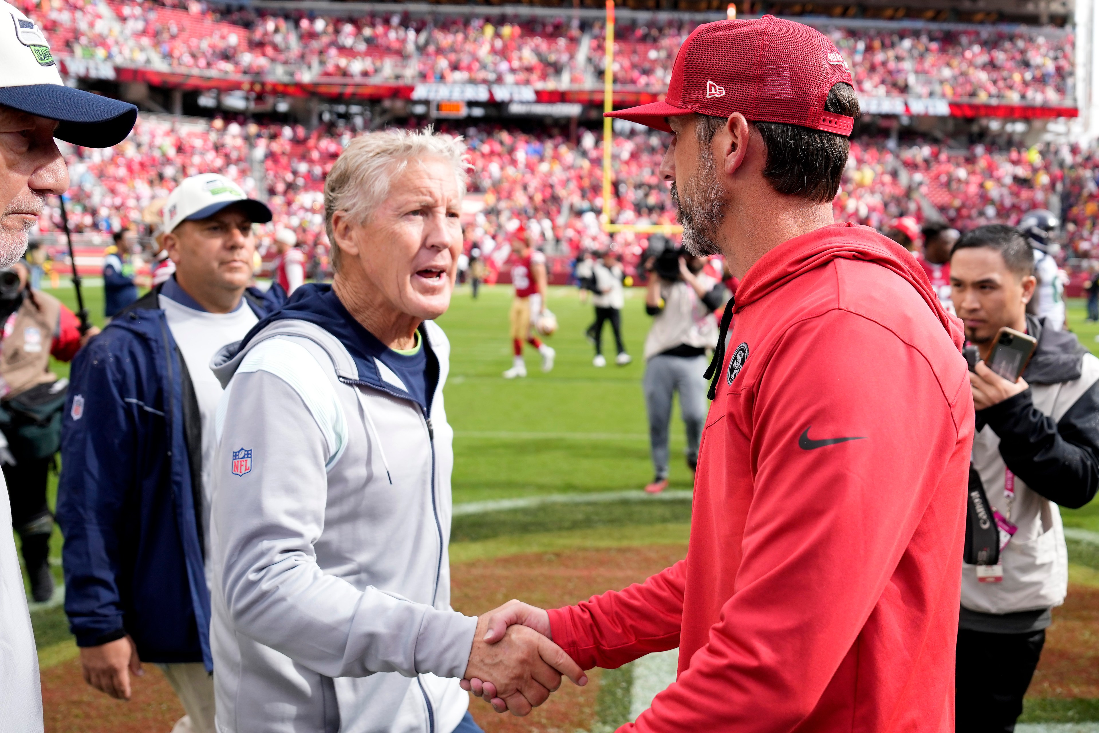 Two men, one in a light jacket and the other in a red hoodie and cap, shake hands on a football field, surrounded by people and a stadium crowd.