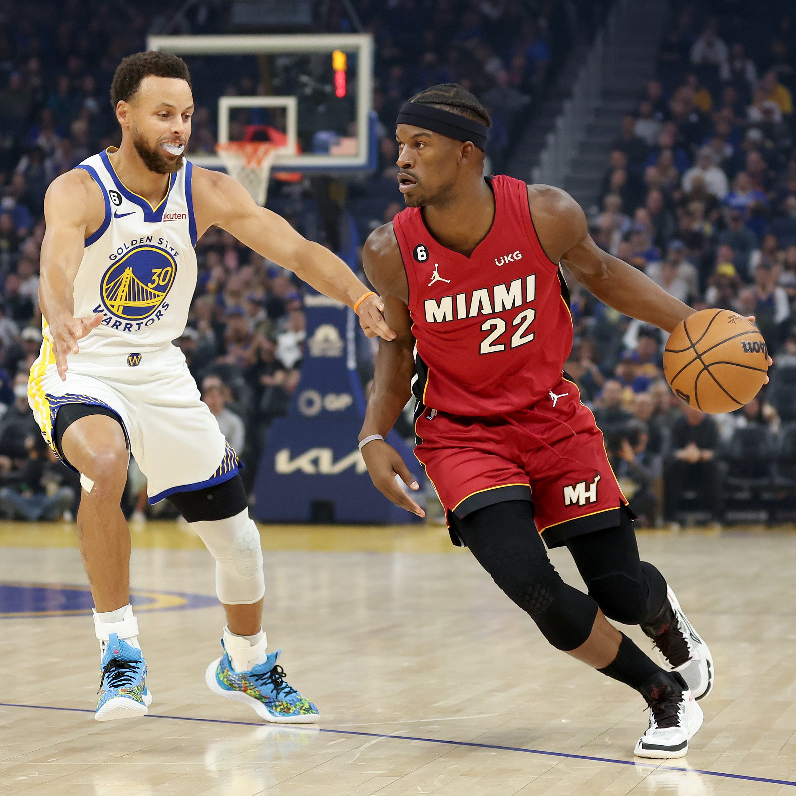 Two basketball players are on a court. One in a Miami red jersey dribbles the ball, while the other in a Golden State white jersey defends closely.