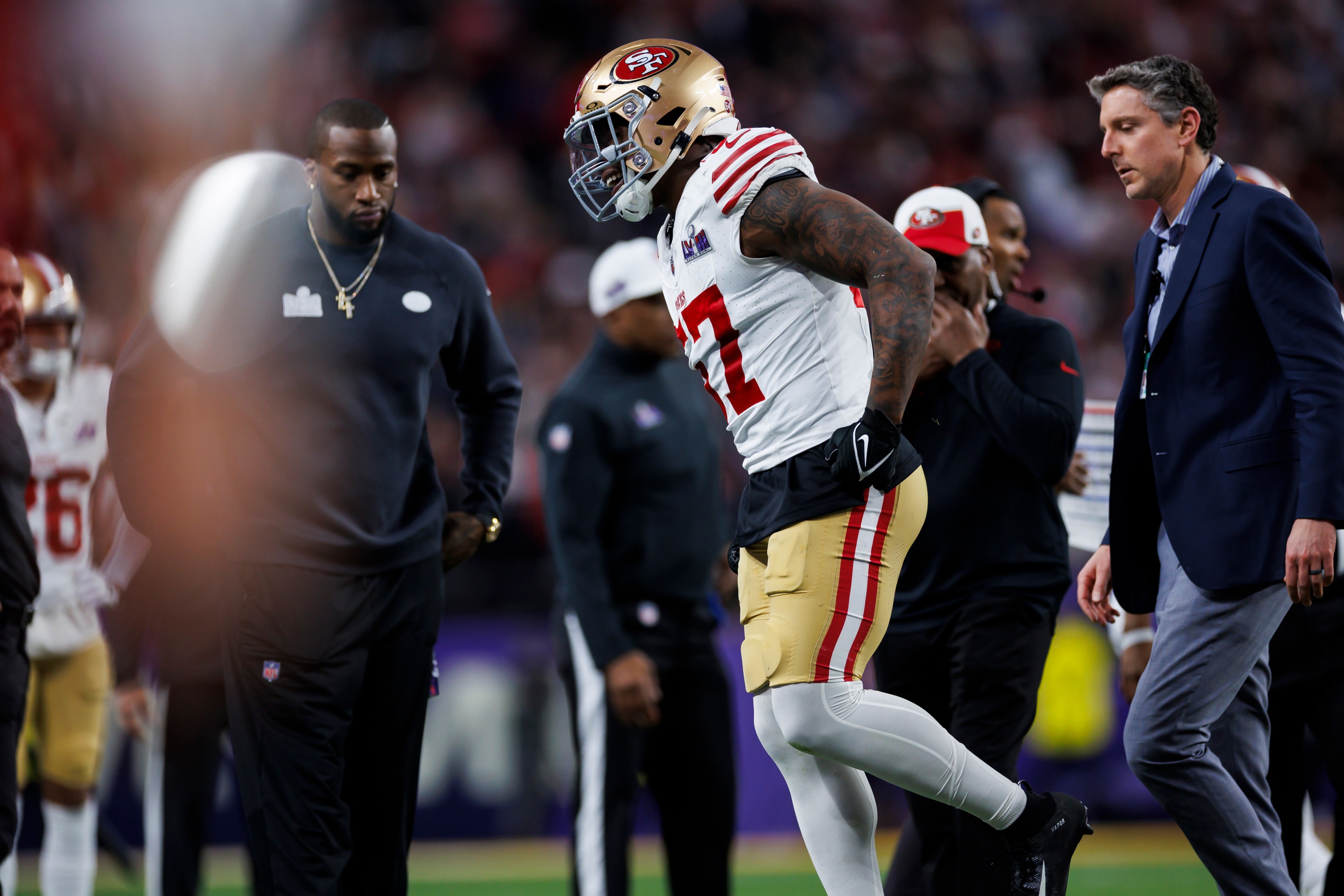 A football player in a 49ers uniform looks distressed, surrounded by two men in casual and formal attire, on a football field with blurred onlookers.