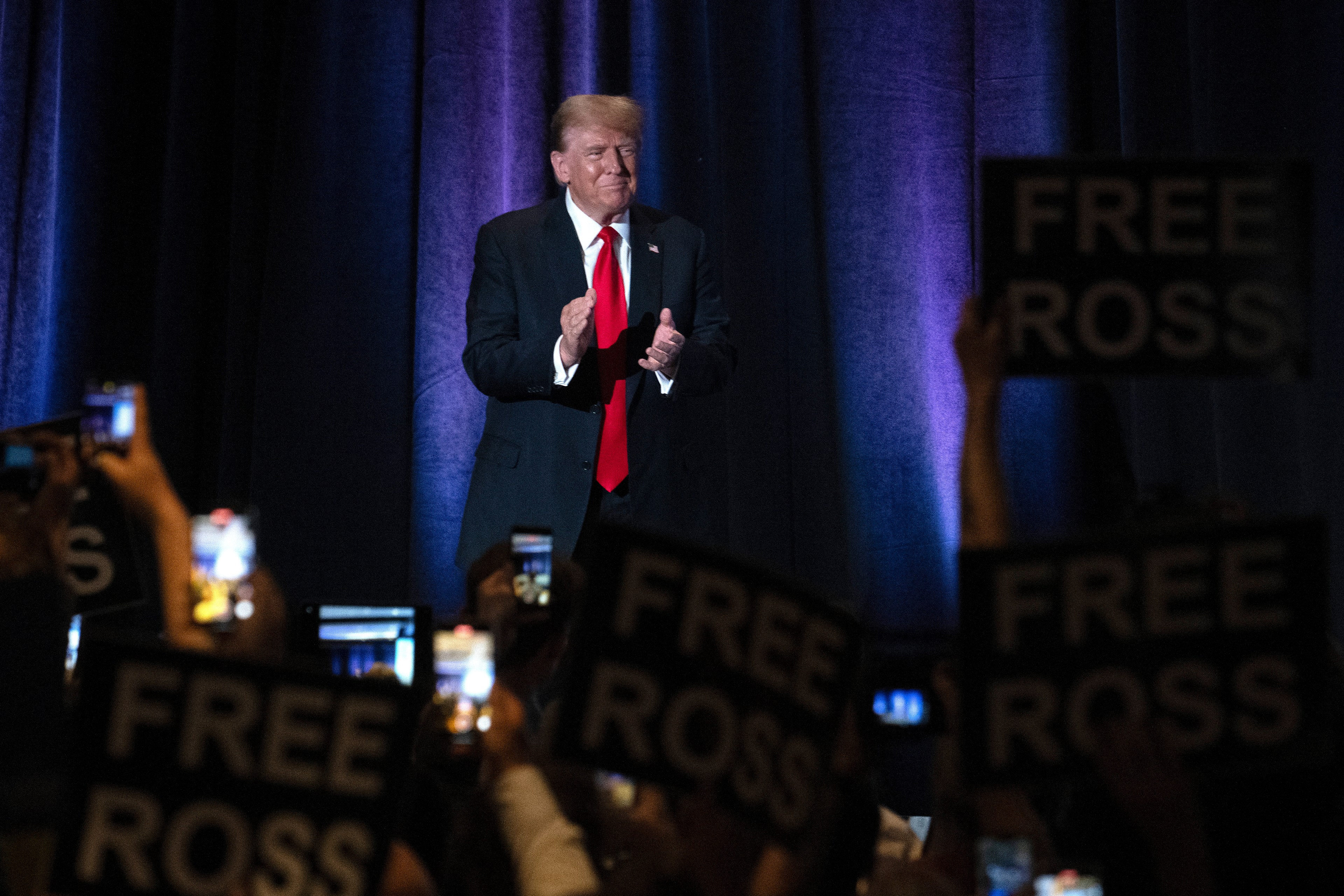 A man in a suit with a red tie claps on stage against a backdrop. The crowd holds signs reading &quot;Free Ross&quot; and takes photos with phones.