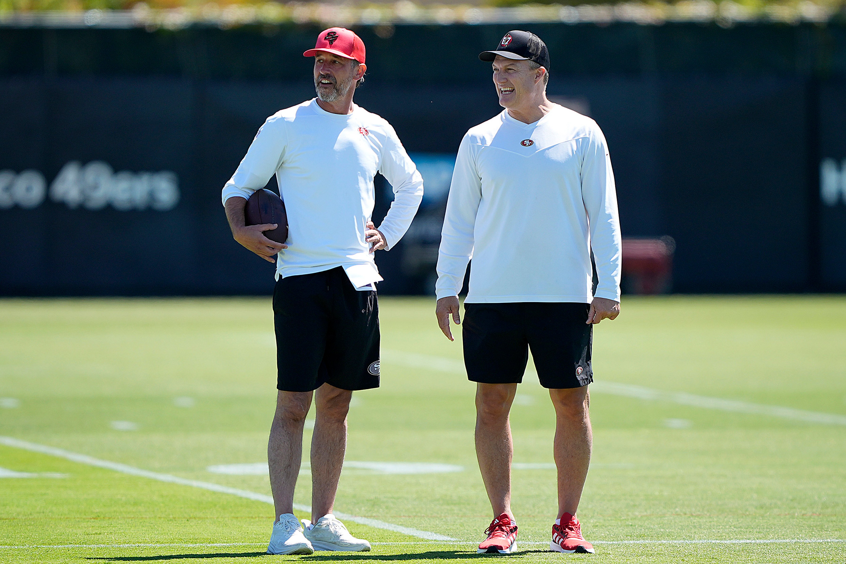 Two men stand on a grassy field, wearing similar sports attire: white long-sleeve tops, black shorts, and caps. One holds a football, and they appear to be smiling.