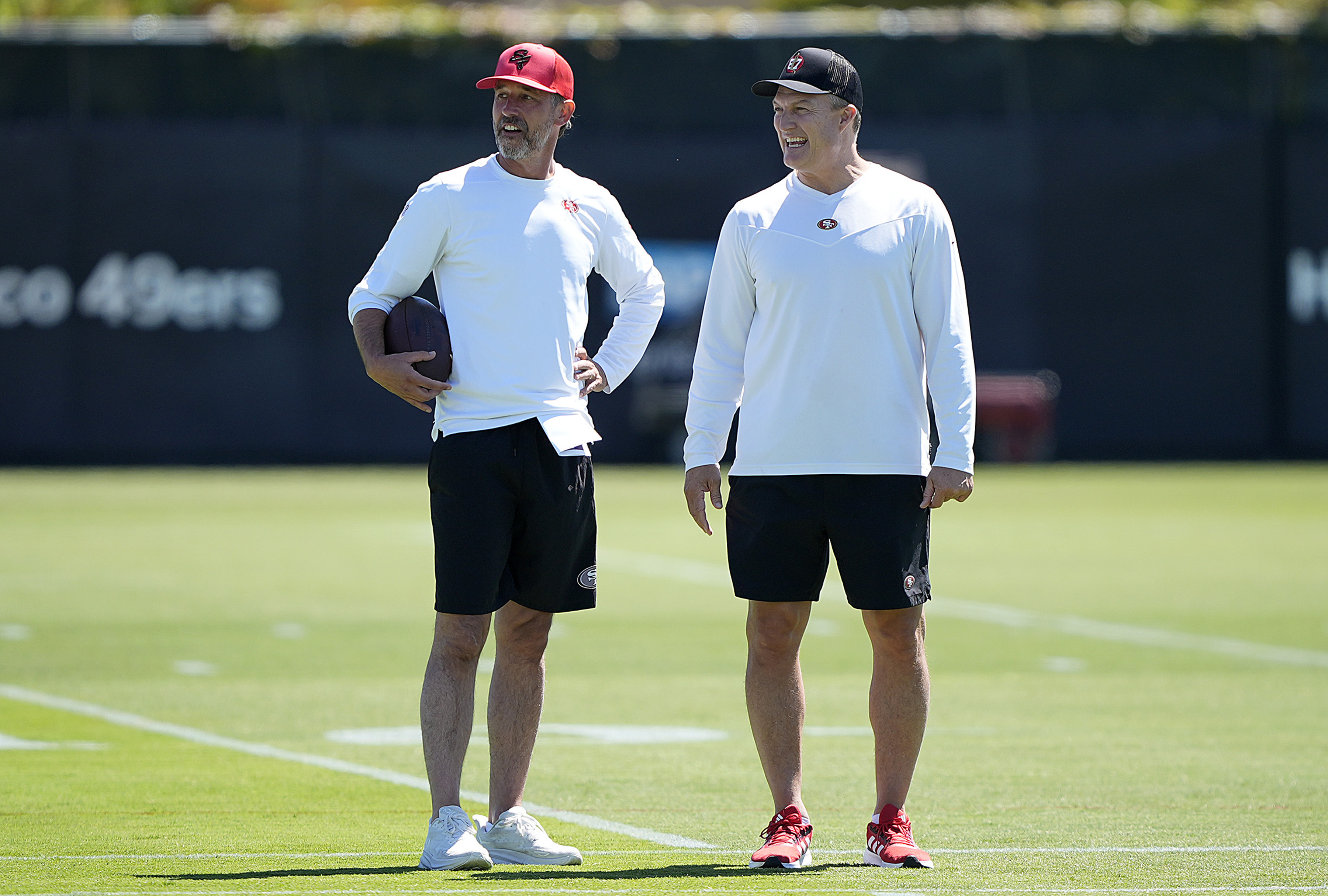 Two men stand on a grassy field, wearing similar sports attire: white long-sleeve tops, black shorts, and caps. One holds a football, and they appear to be smiling.