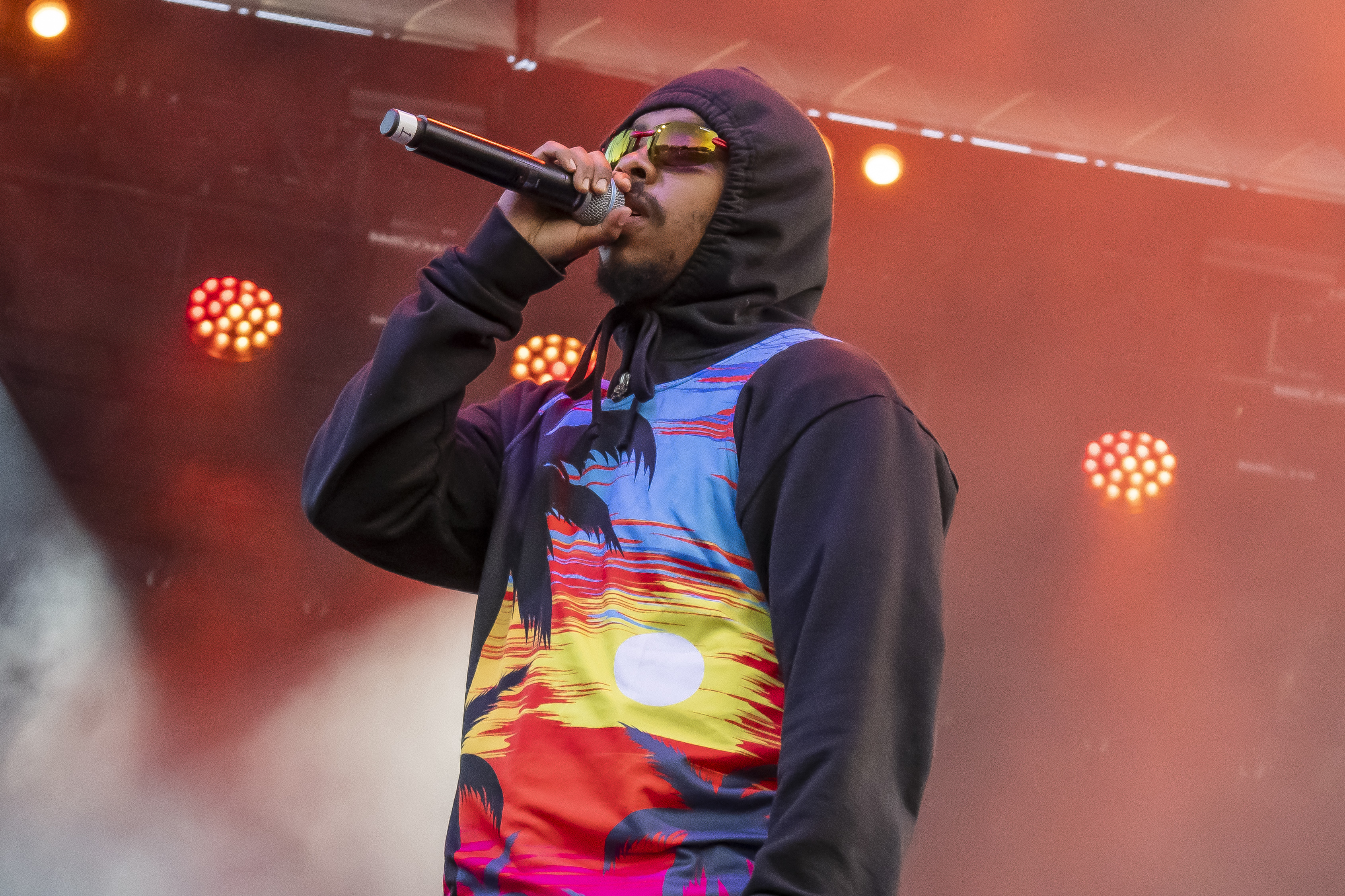 A person is performing on stage, wearing a colorful hoodie with a tropical sunset design. They hold a microphone and wear sunglasses, with stage lights behind them.