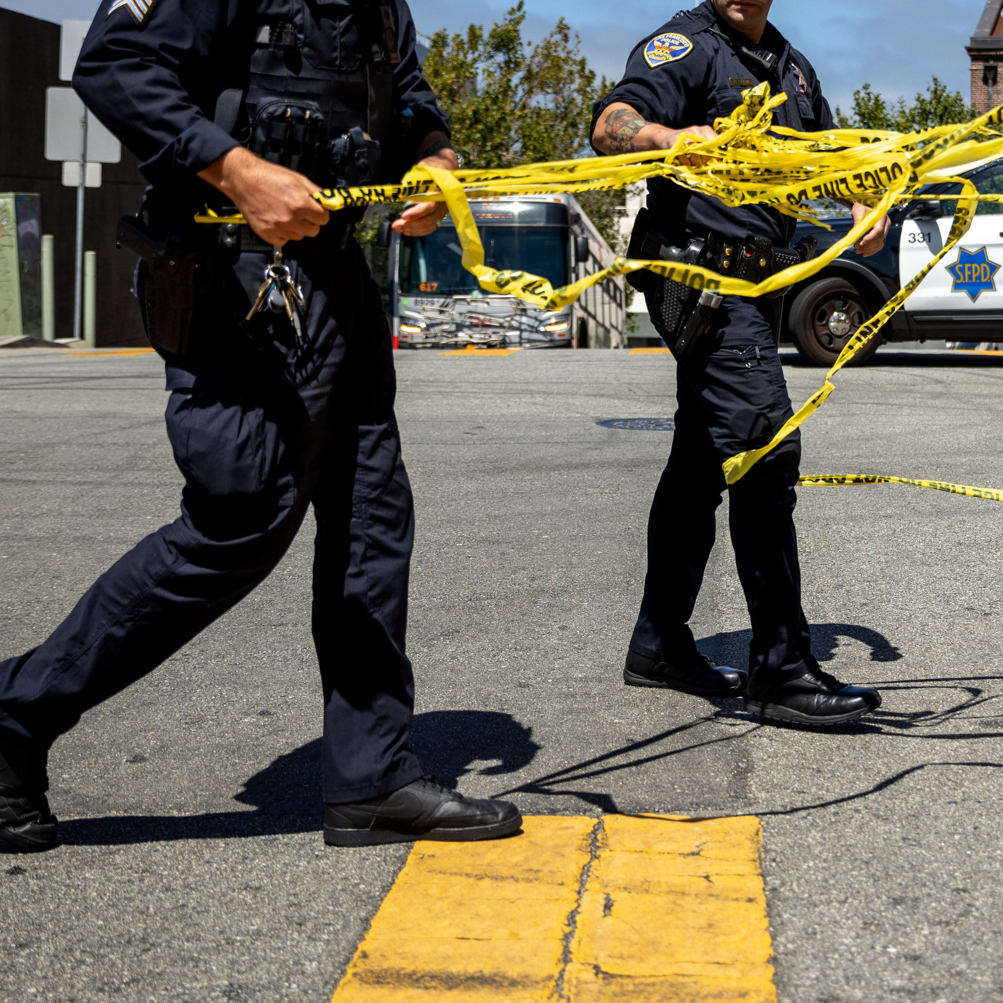 Two police officers are laying out yellow caution tape at a street intersection. A police car is parked nearby, and a bus is visible in the background.