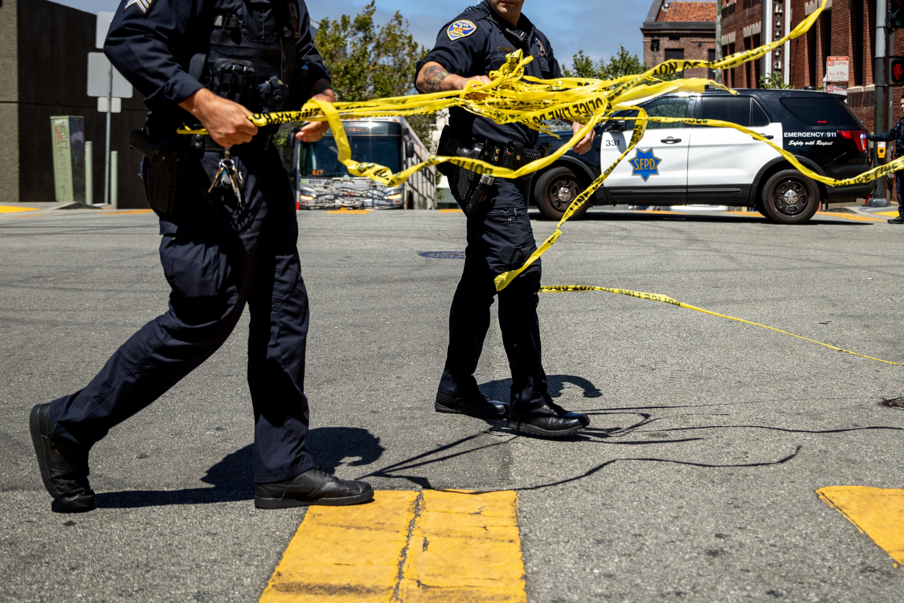 Two police officers are laying out yellow caution tape at a street intersection. A police car is parked nearby, and a bus is visible in the background.